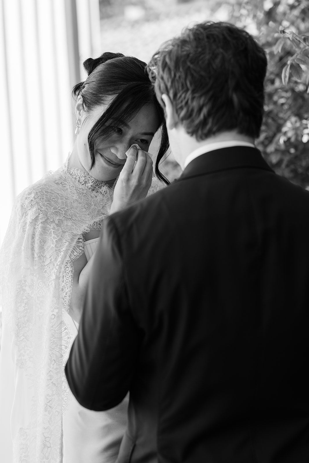 A tender black and white wedding moment shows a bride in lace wiping away tears while facing her groom.