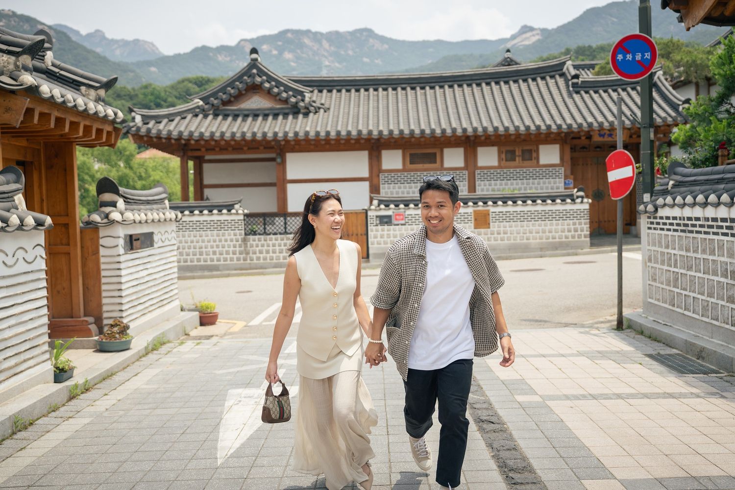 Visitors walk along stone pathways between traditional Korean palace buildings with mountains visible in the background.