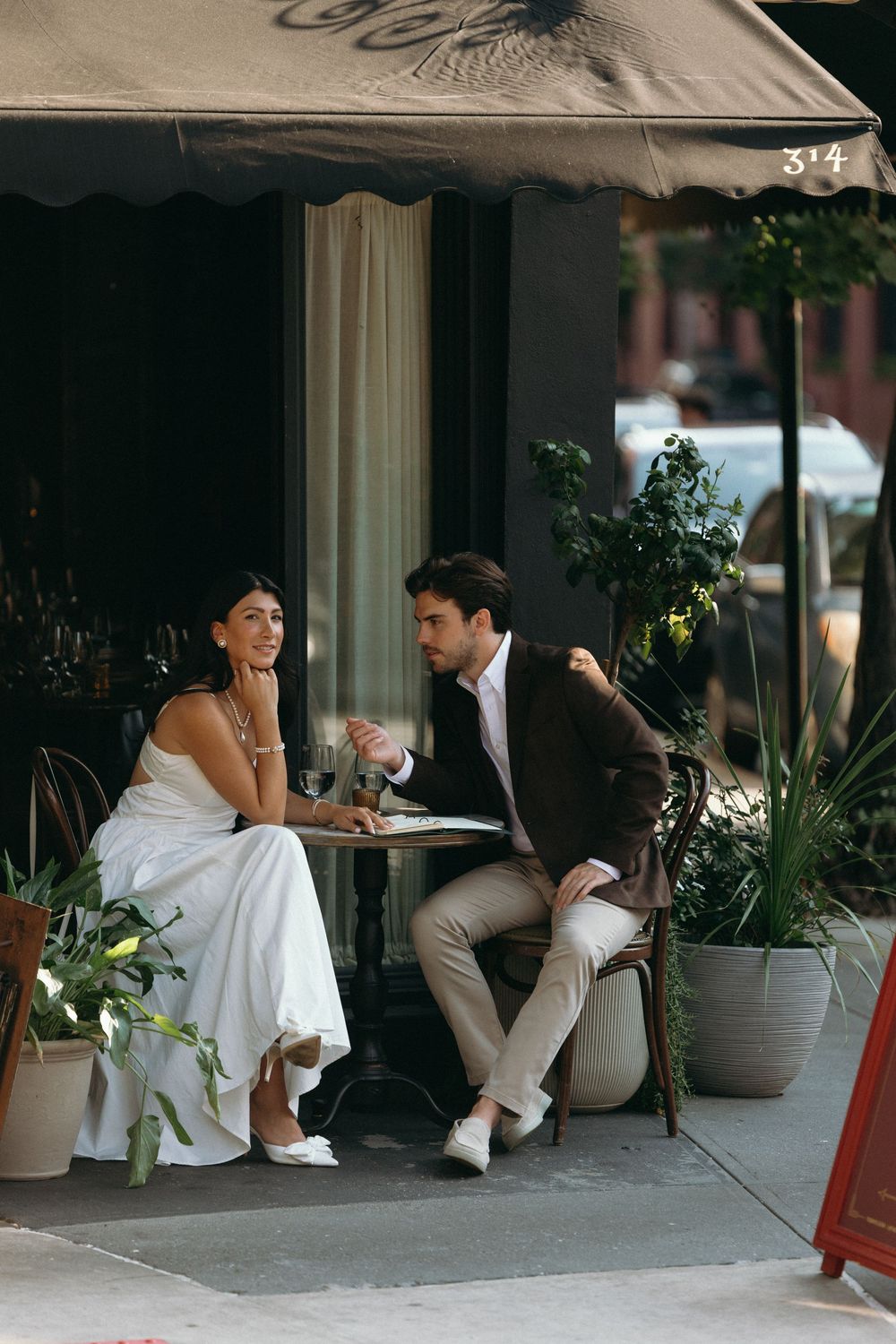 A couple sits together on a bench outside a building with white planters and greenery in a candid sequence of photos.
