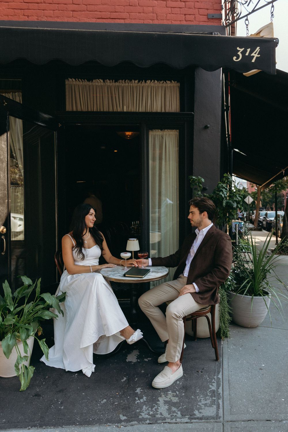 A couple sits together at a sidewalk cafe table in front of a brick building with storefront windows.