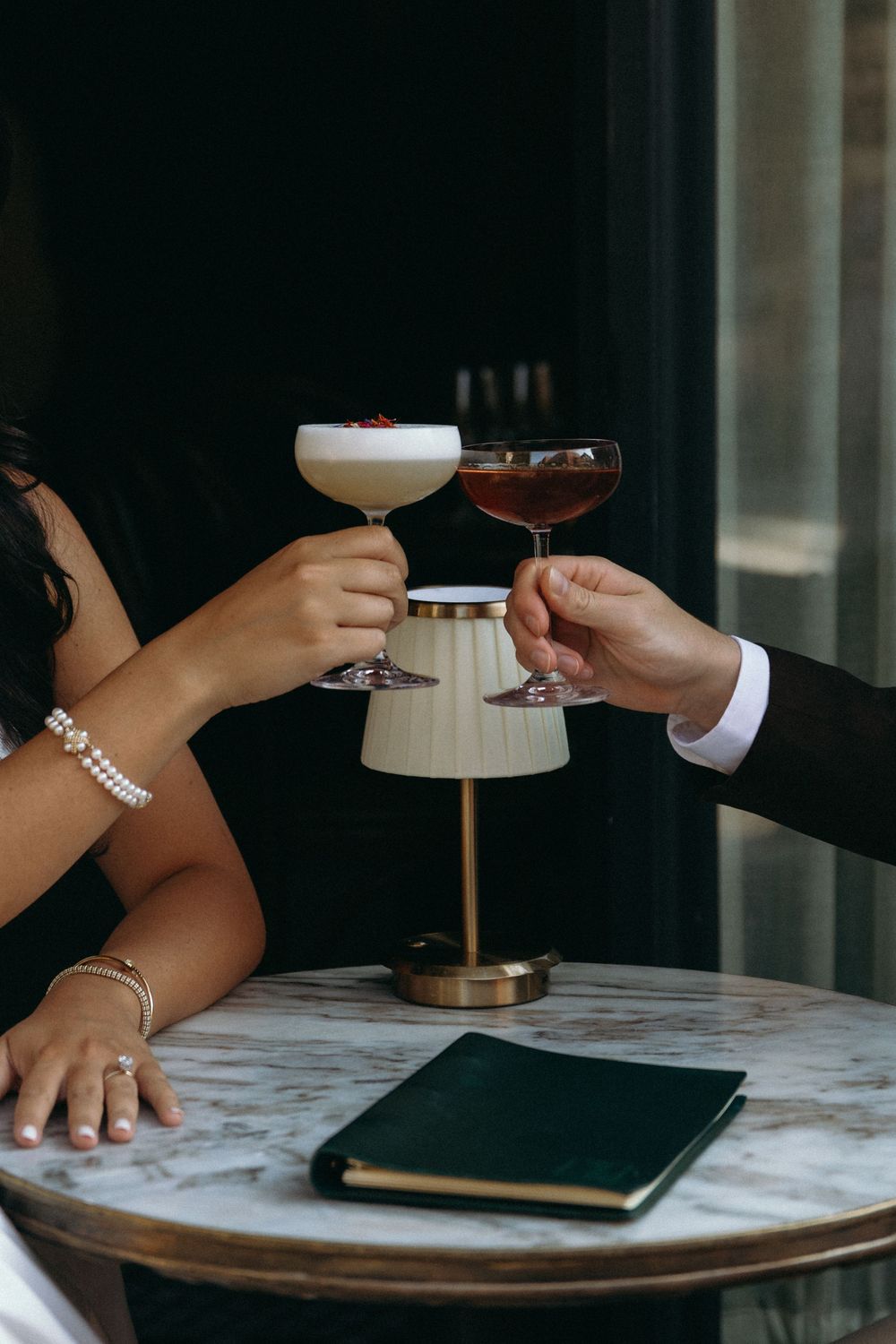 Two people holding cocktail glasses over a marble table with a black notebook during an elegant evening meeting.
