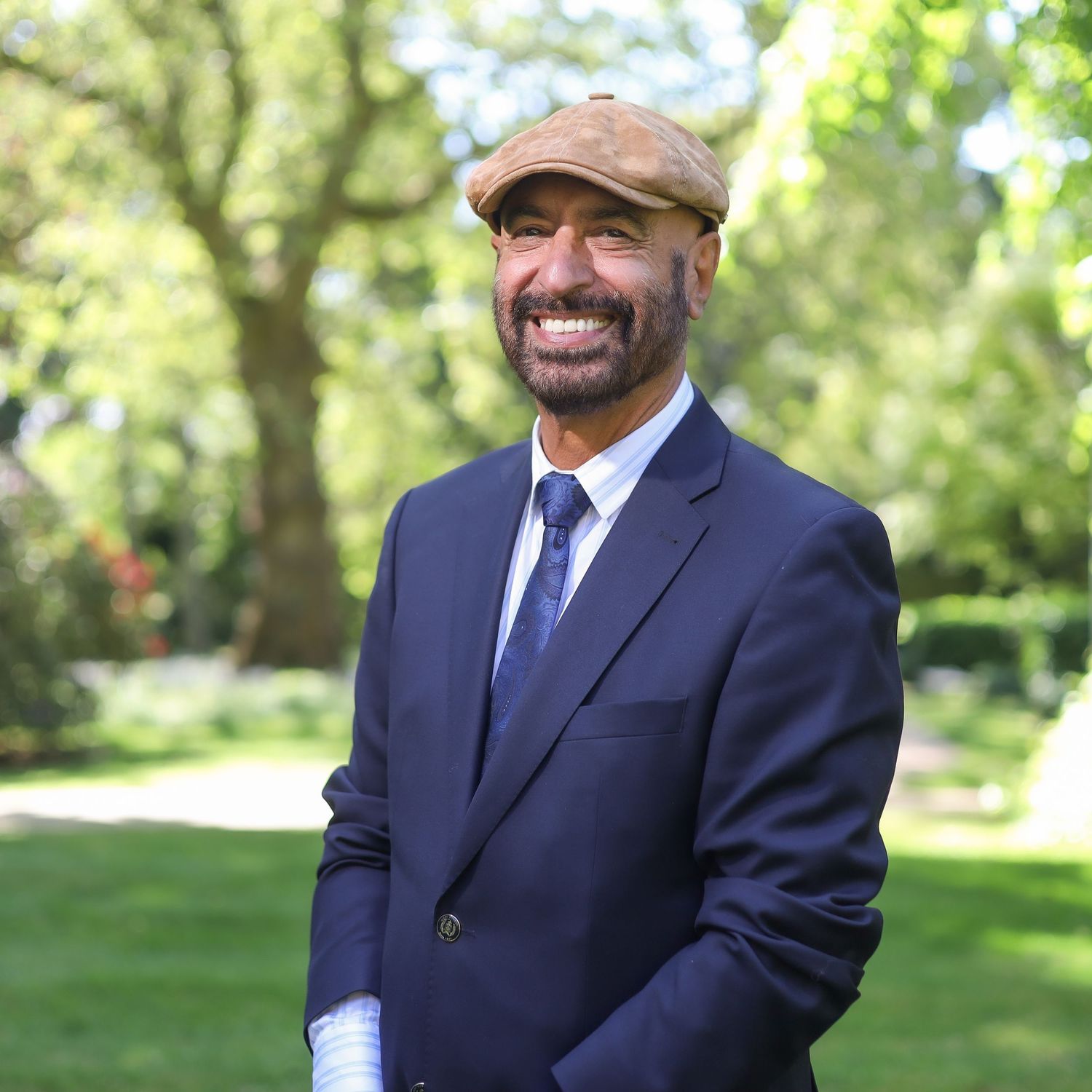 Professional wearing a navy suit and flat cap smiles confidently in a sunny outdoor garden setting.