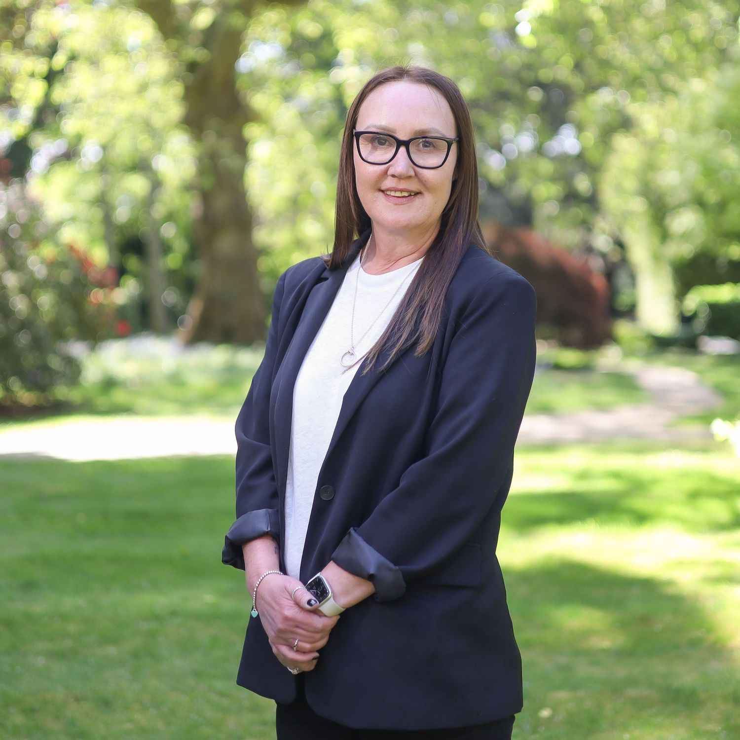 Professional portrait in black blazer and white top with glasses in garden setting.