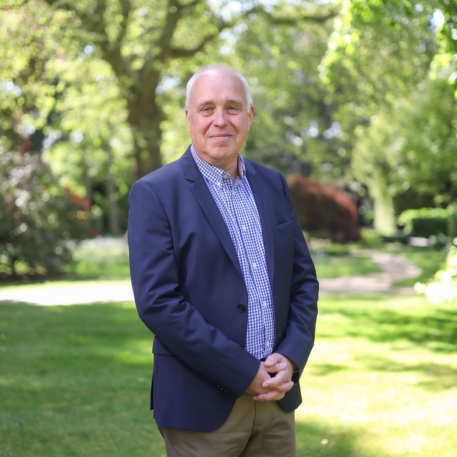 Professional in navy suit jacket and striped shirt poses in front of trees in outdoor garden setting.