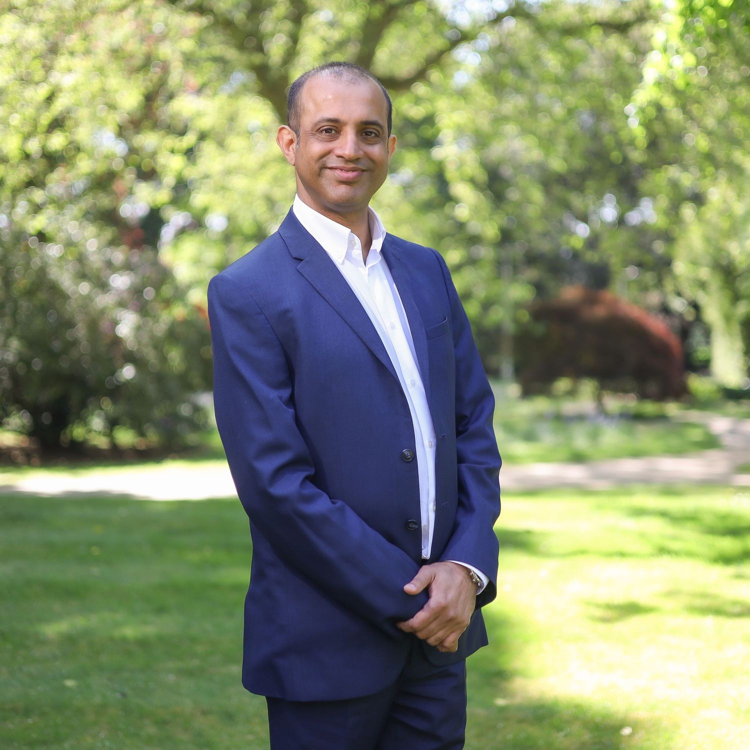 Professional in a navy jacket stands outdoors in a sunny park setting with green trees and grass in the background.