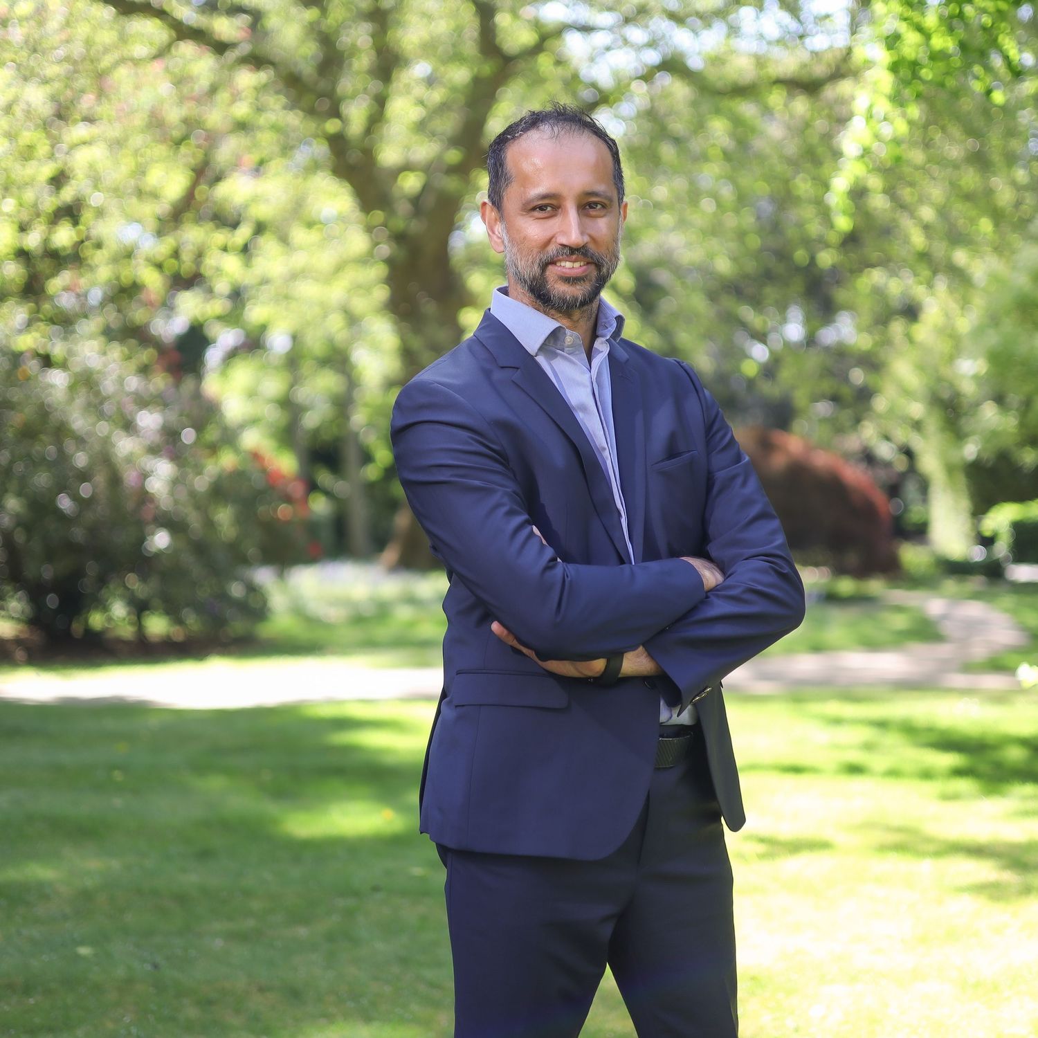 Person in a dark business suit poses with crossed arms against a lush green park background.