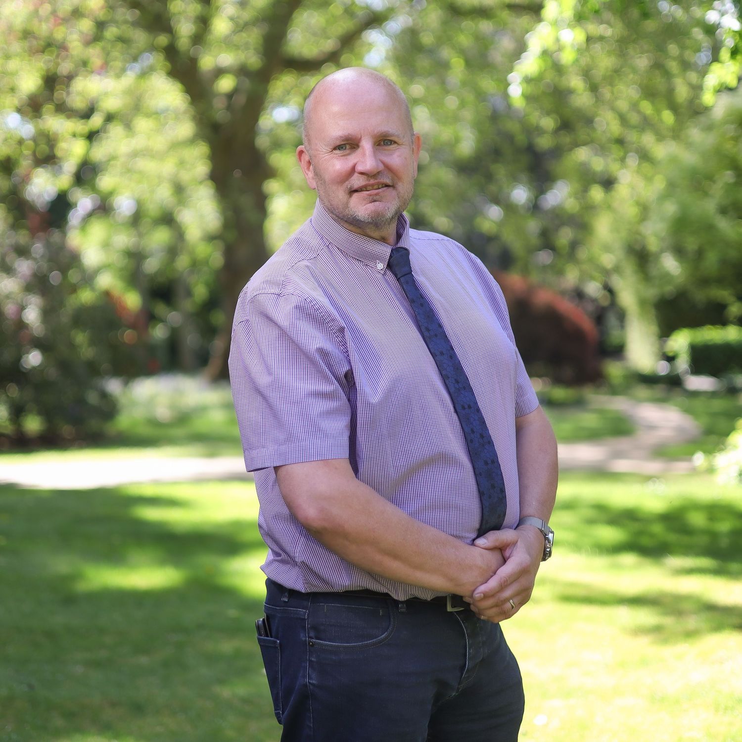 A person wearing a purple striped shirt and dark pants poses in a sunny outdoor park setting.
