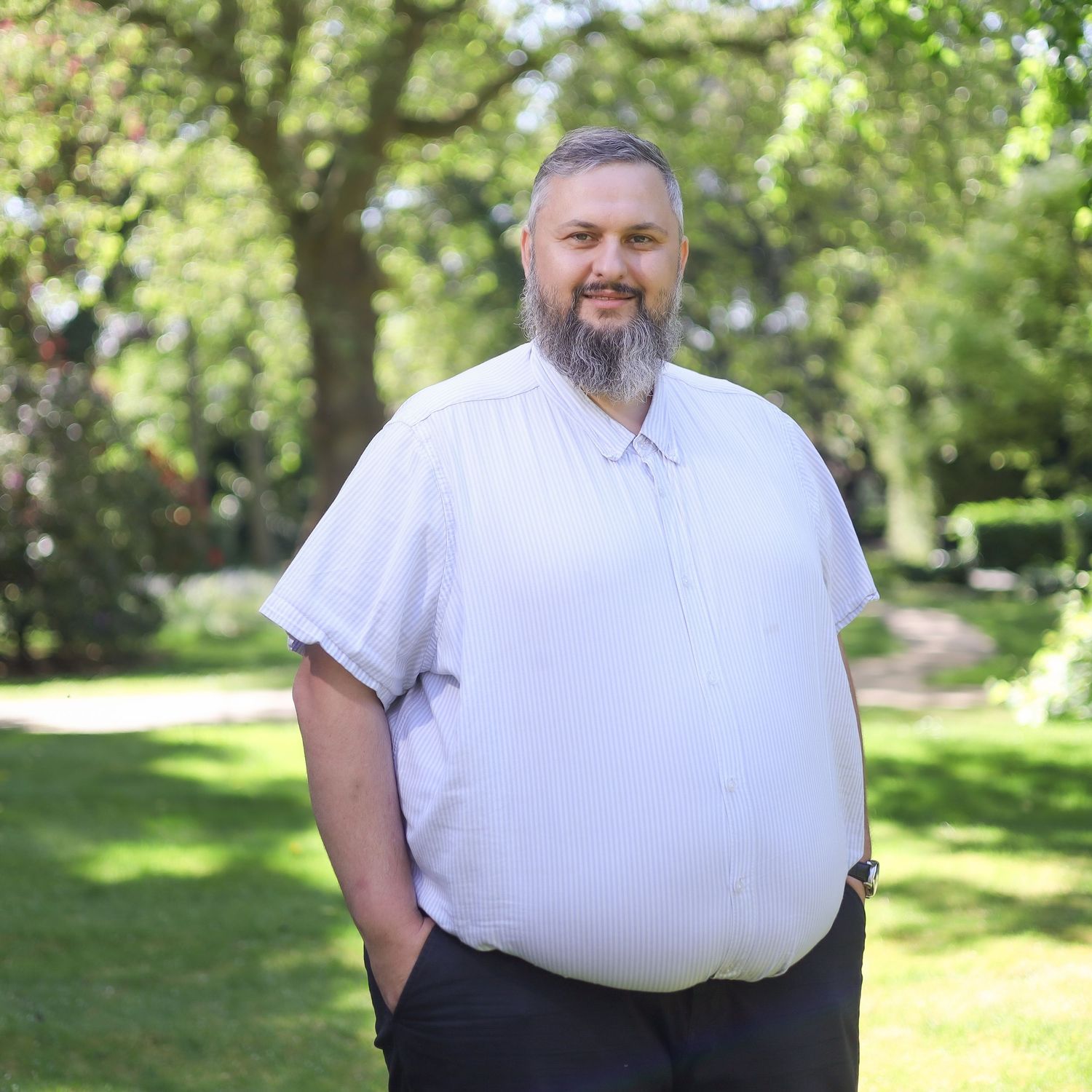 Individual in a white dress shirt and dark pants stands casually in a park setting.