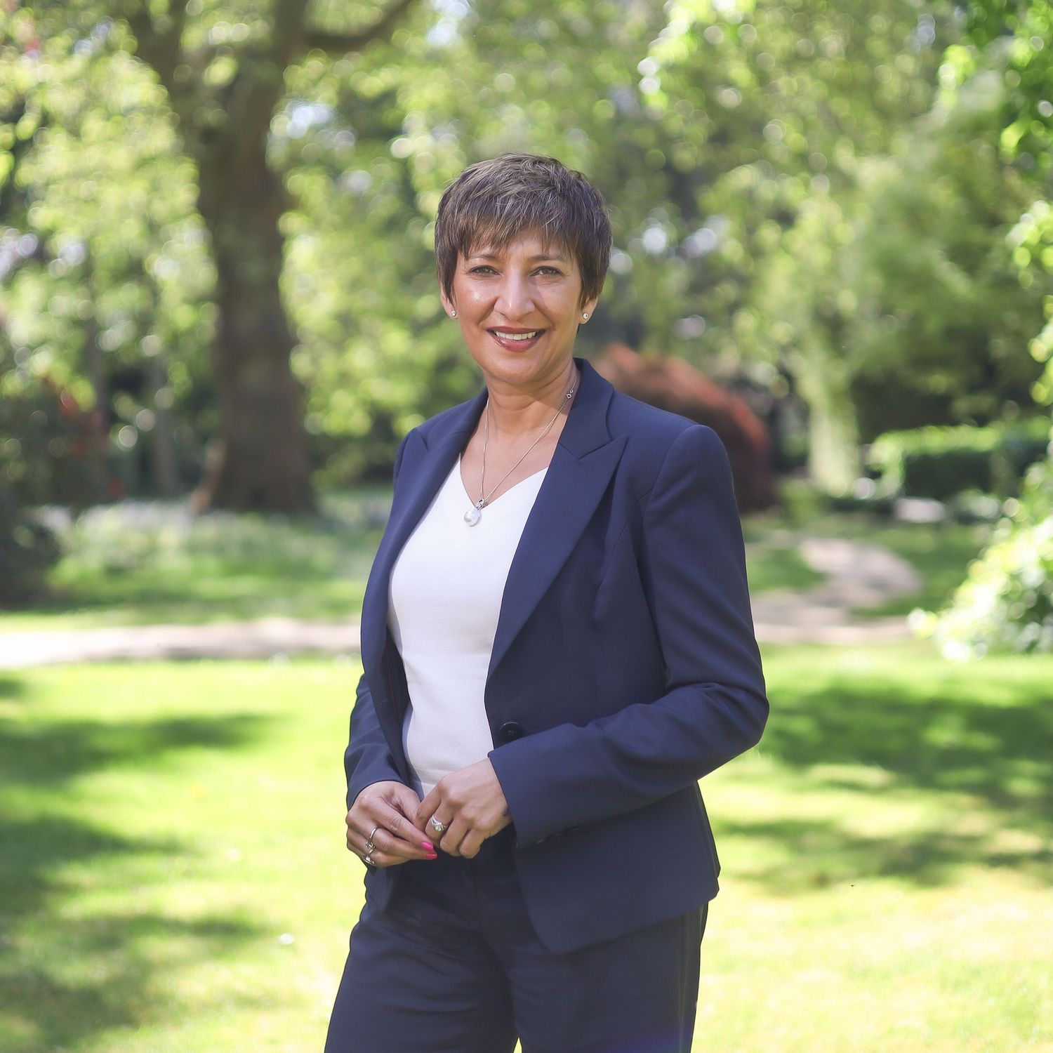 A person wearing a navy blazer and white top poses in a sunny garden setting.