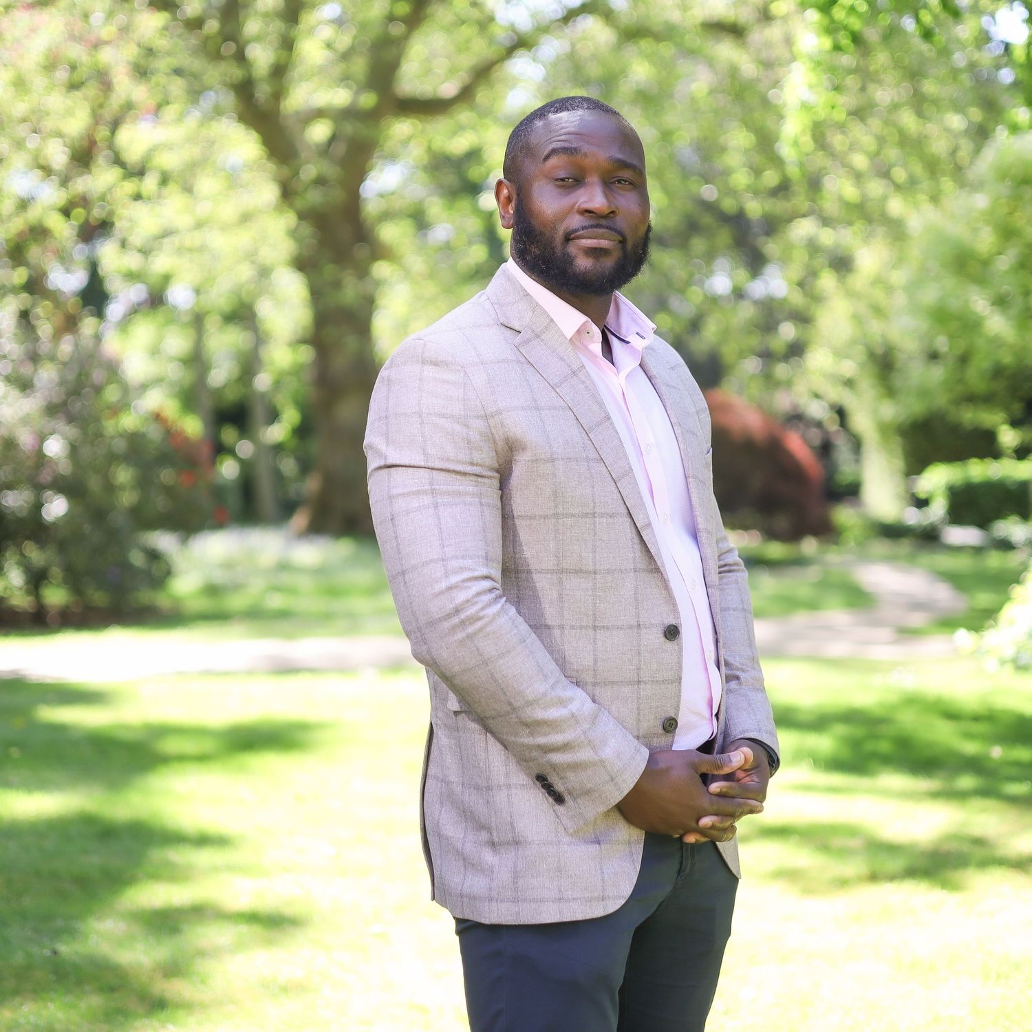 Professional in light checkered blazer poses confidently in outdoor park setting with green foliage backdrop.