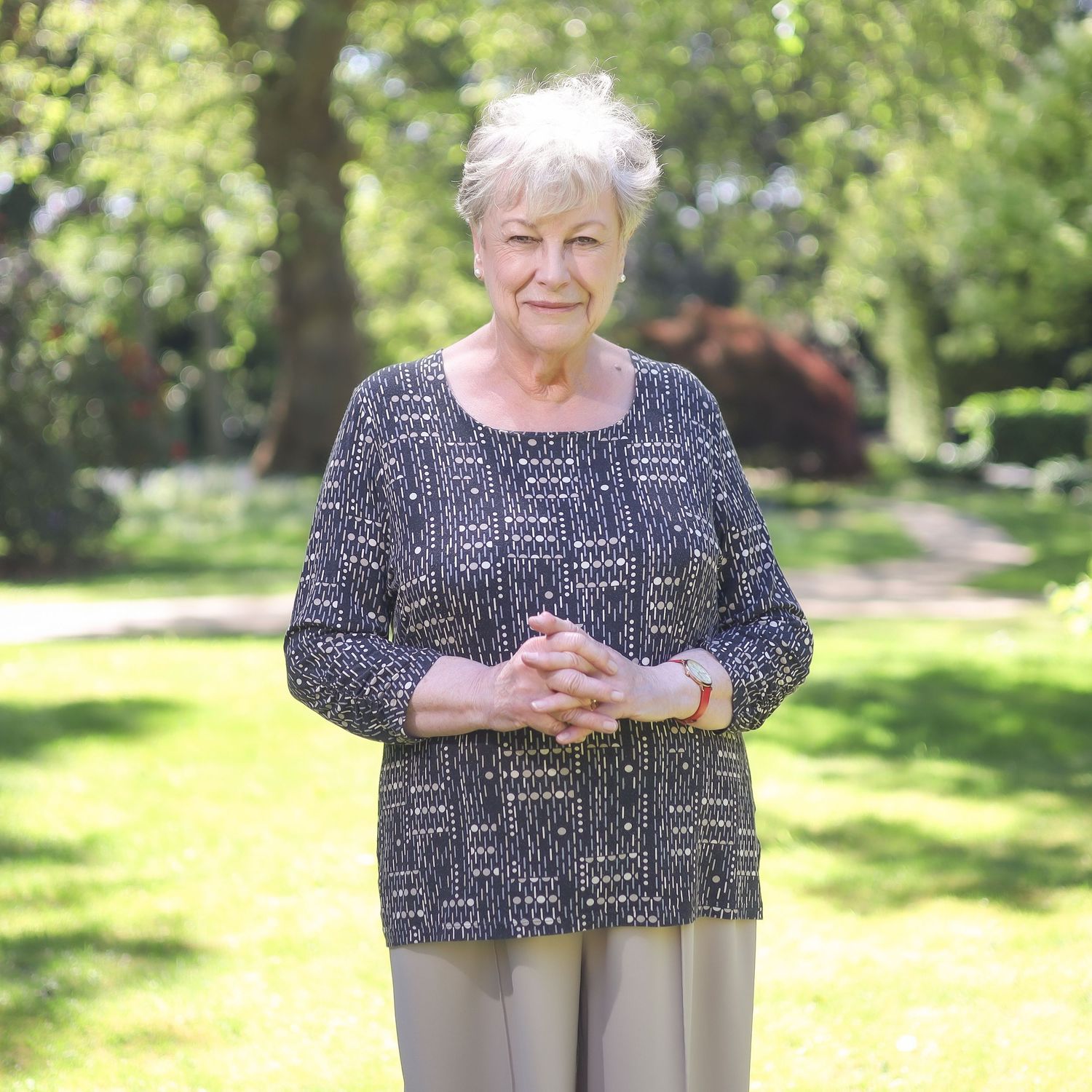 Person in a patterned top and gray pants stands outdoors in a sunny garden environment.