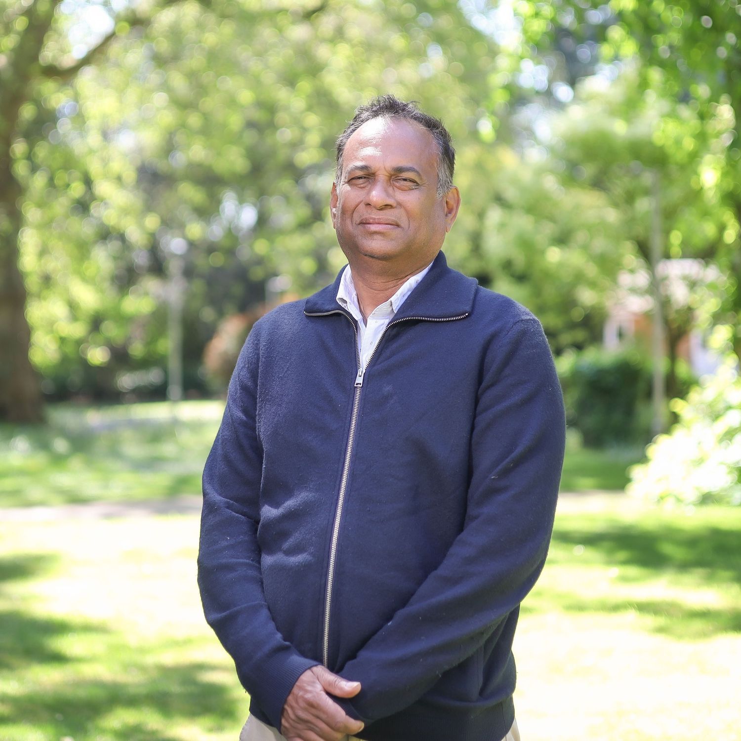 Person wearing a navy blue zip-up jacket poses casually against a bright natural outdoor garden backdrop.