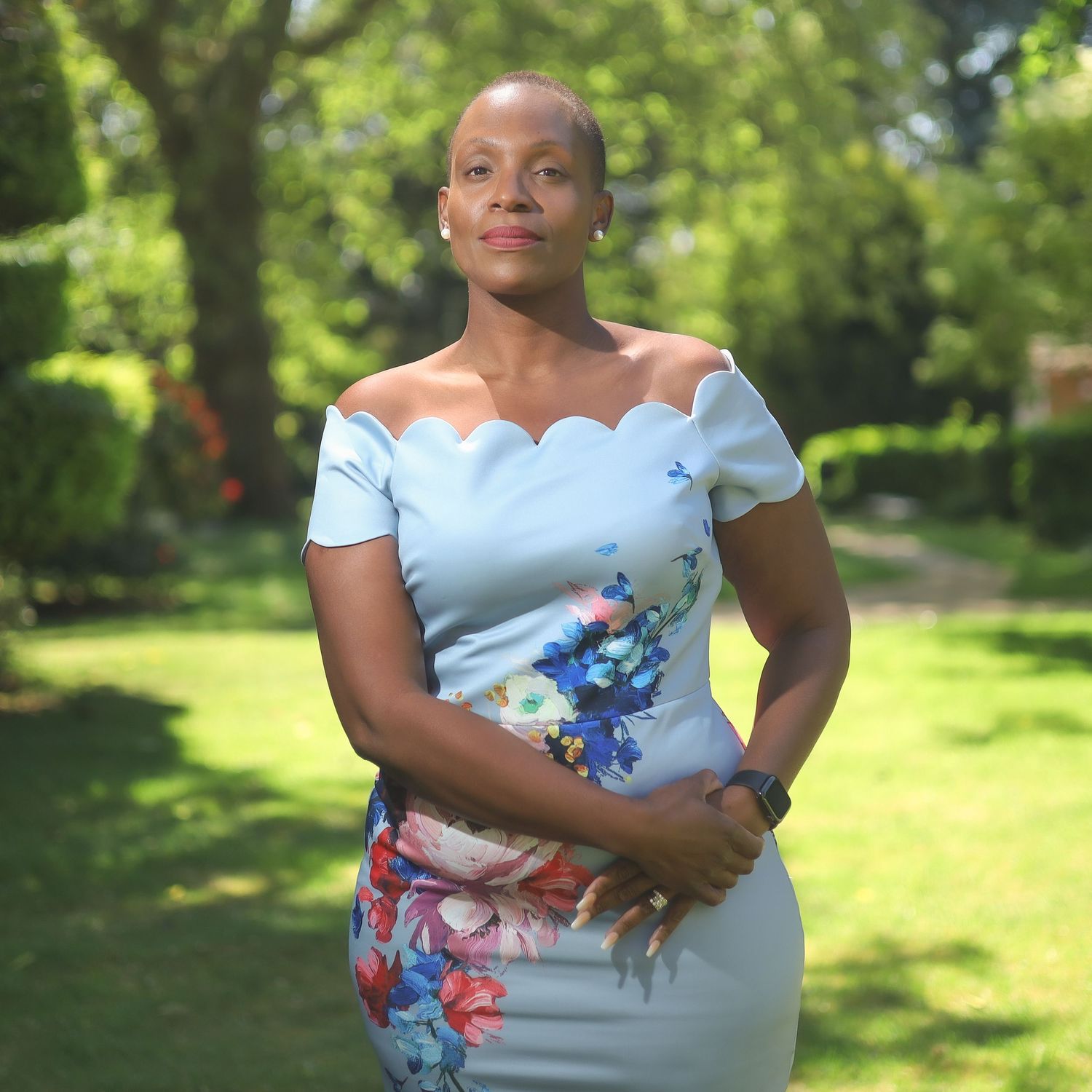 Woman in a light blue floral dress poses elegantly in a sunlit garden setting.