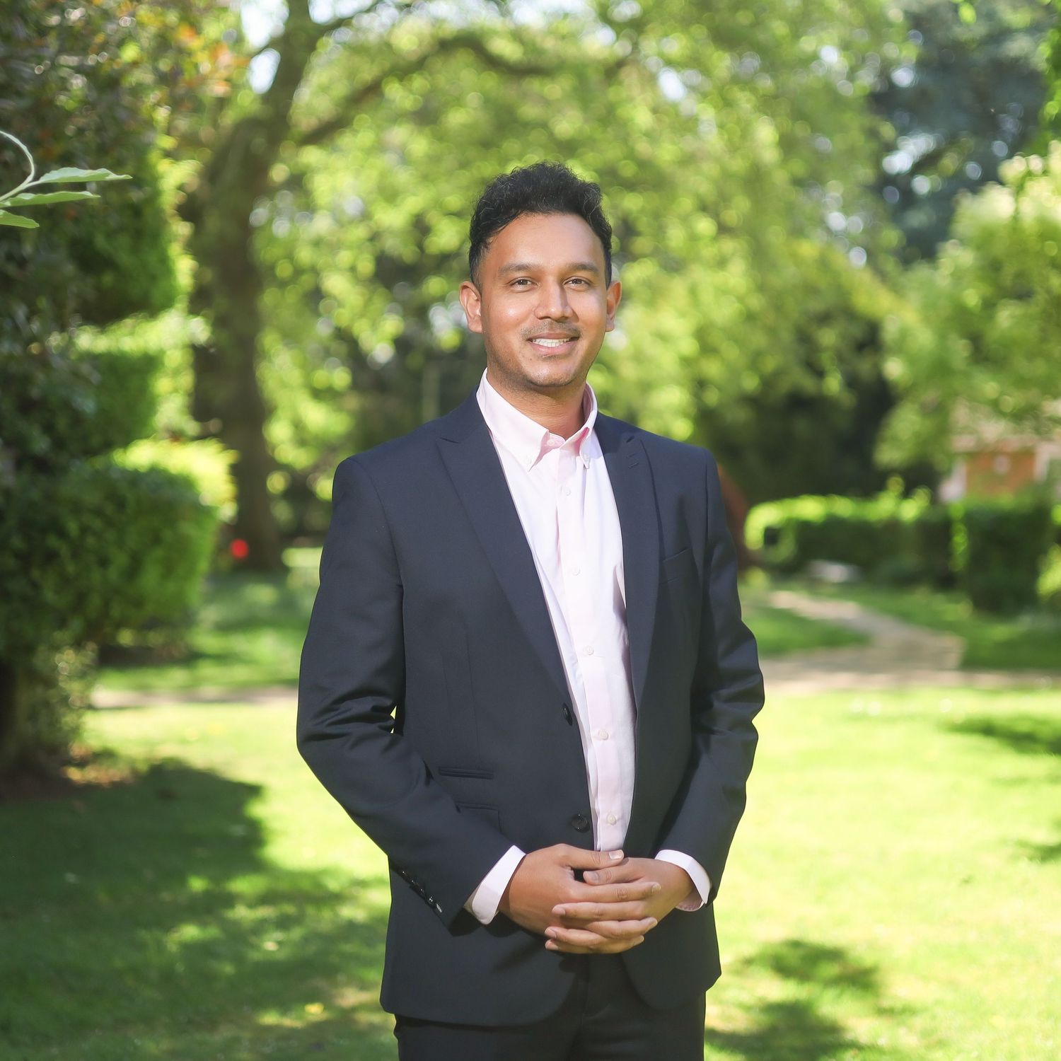 Professional headshots in dark suit and white shirt with sunny garden backdrop.