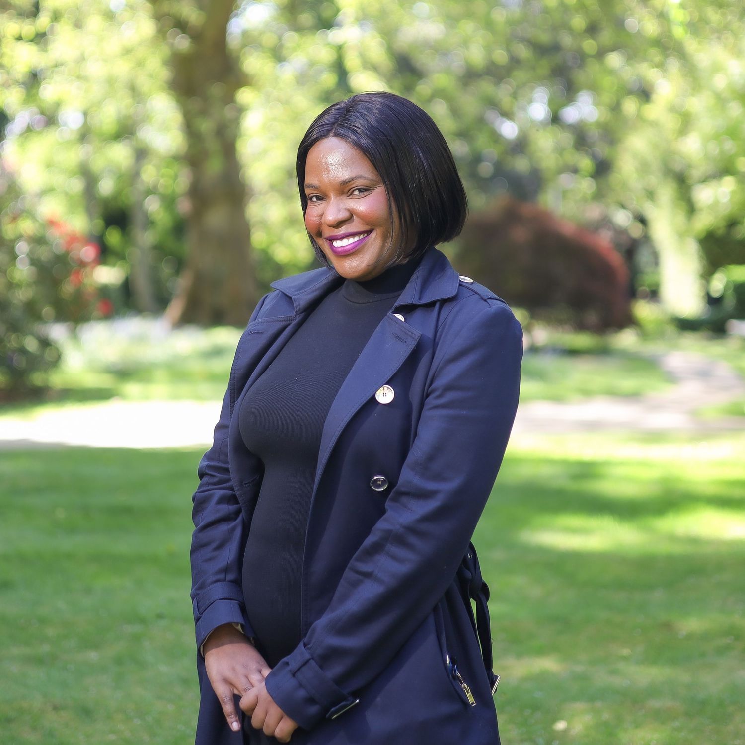 Professional wearing a navy blazer and black top stands smiling outdoors in a sunny garden setting.