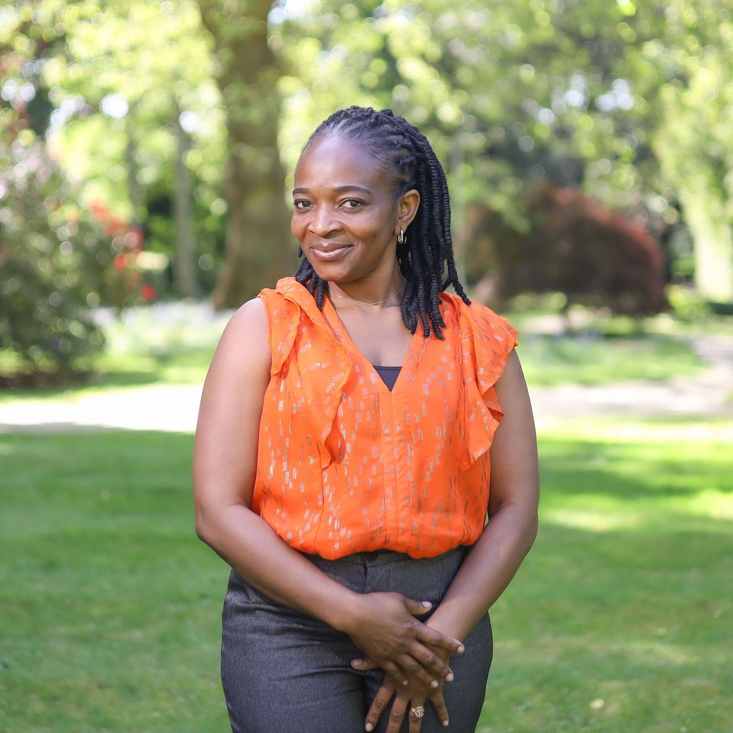 A person in a bright orange sleeveless top poses outdoors in a sunny garden setting.