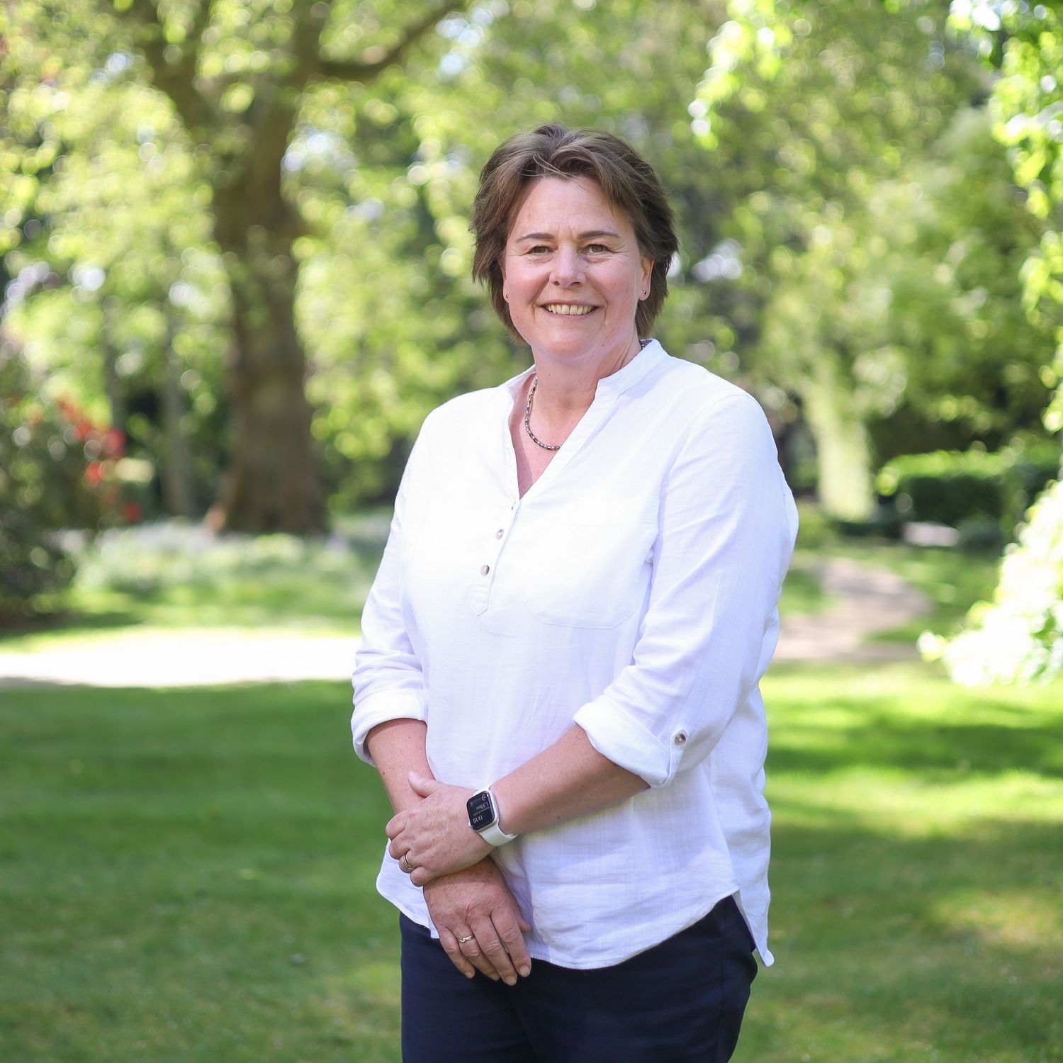 Professional in white shirt stands outdoors in a sunny garden setting with lush green trees in background.