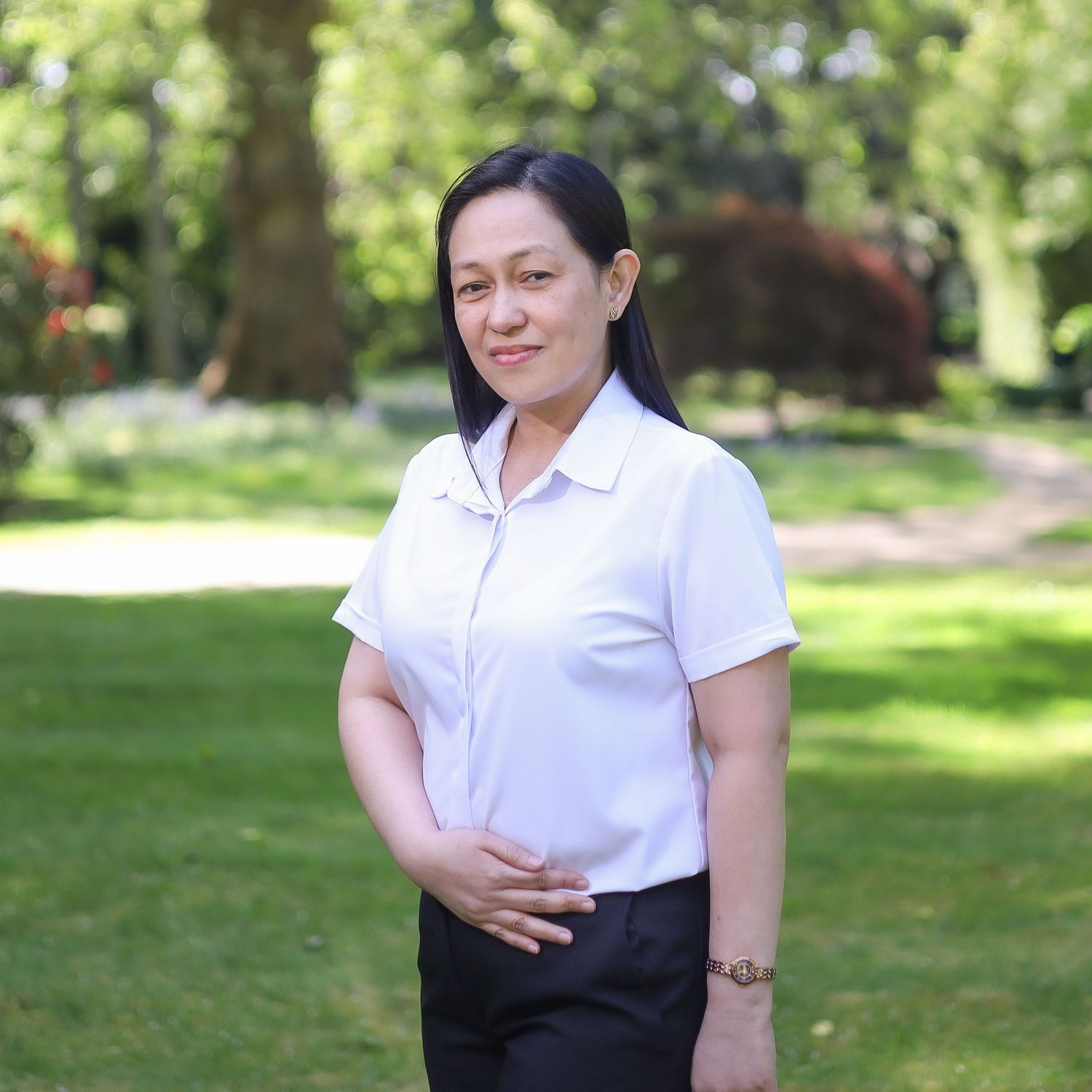 Person in white blouse and dark skirt standing outdoors in a sunny garden setting with green foliage background.