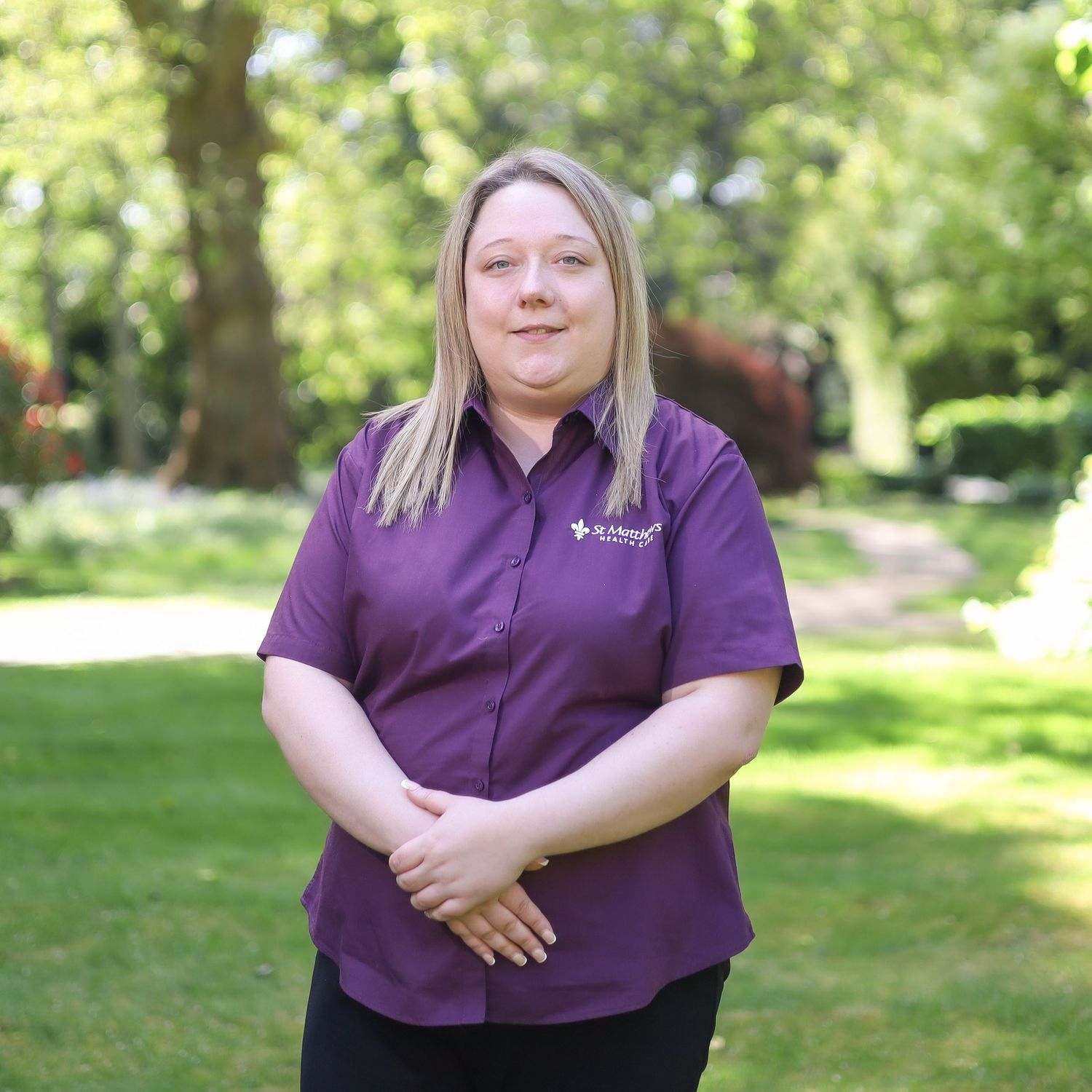 A person wearing a purple polo shirt stands in front of lush green trees and grass.
