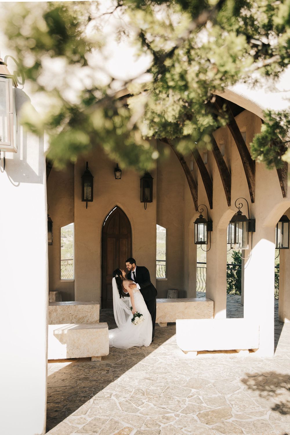 A couple stands in a romantic Mediterranean-style colonnade with white arches and hanging lanterns.