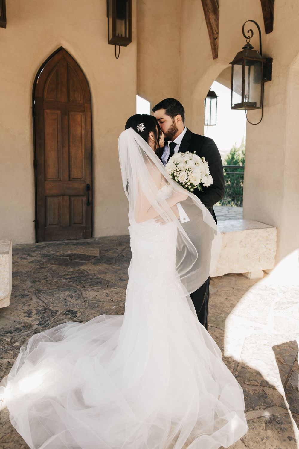 Romantic wedding couple embraces in archway with flowing white dress and lanterns at elegant stone venue.