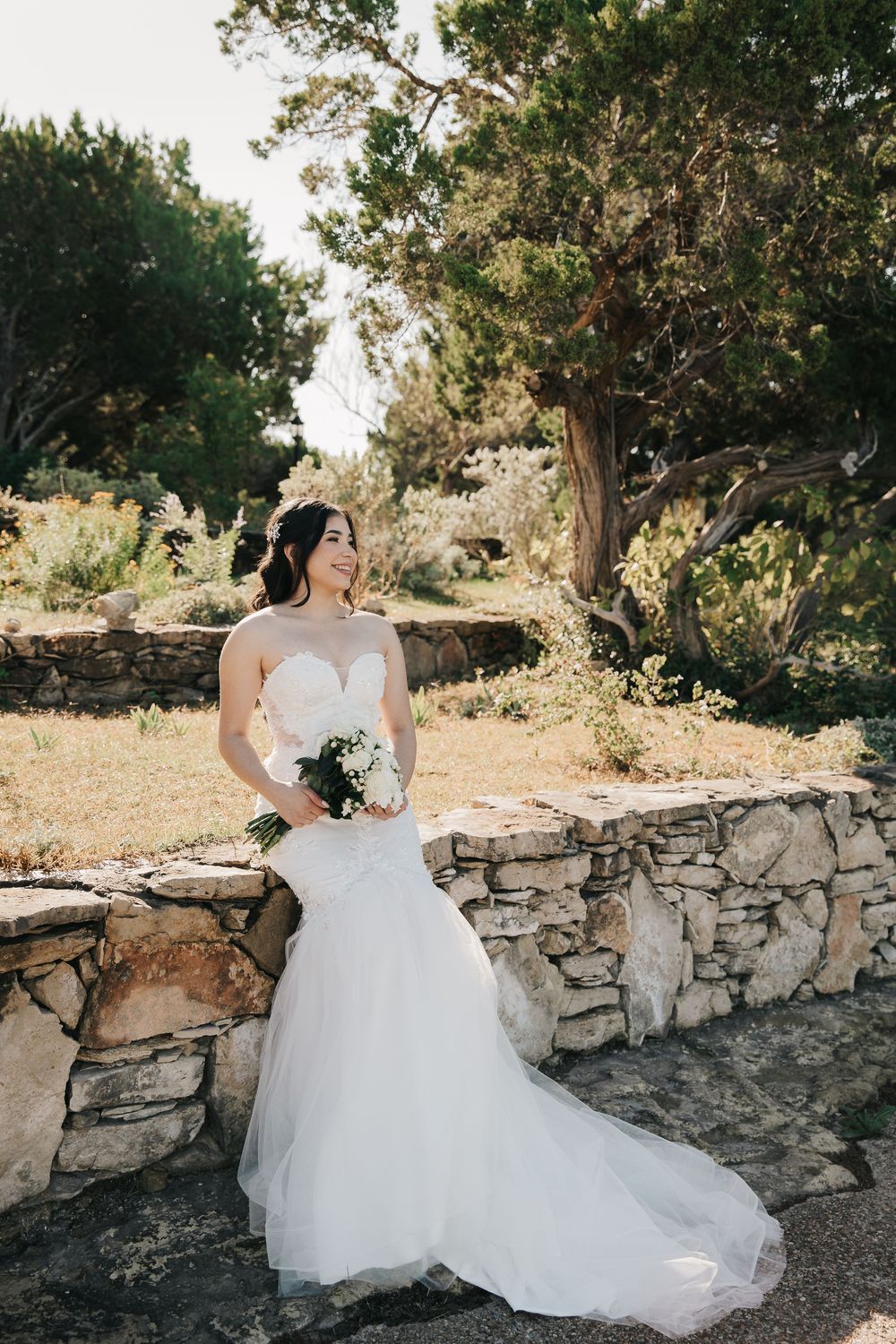 Bride in a flowing white wedding dress sits on rustic stone wall surrounded by trees in natural outdoor setting.