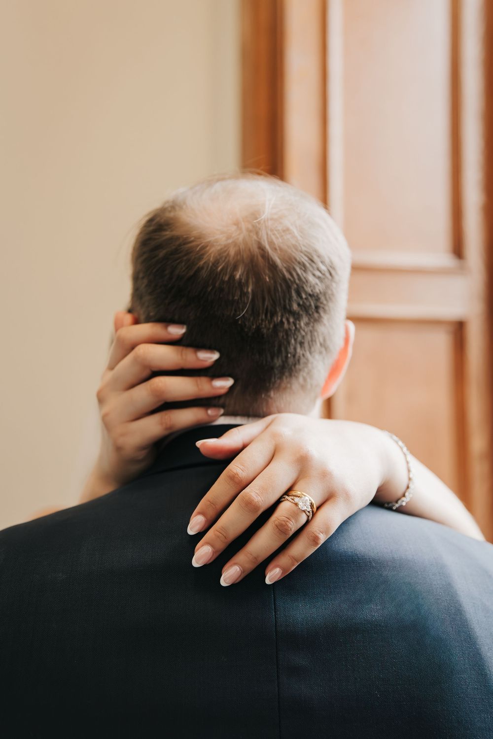 A hand with a wedding ring rests gently on the back of a navy blue suit jacket in warm lighting.