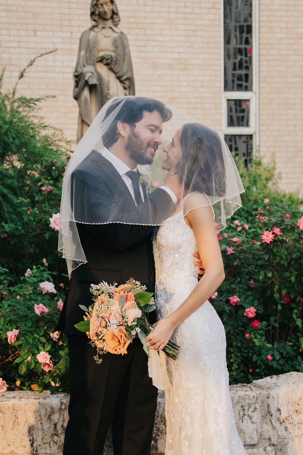 A romantic wedding couple shares an intimate moment in front of a church with blooming rose bushes and stone architecture.