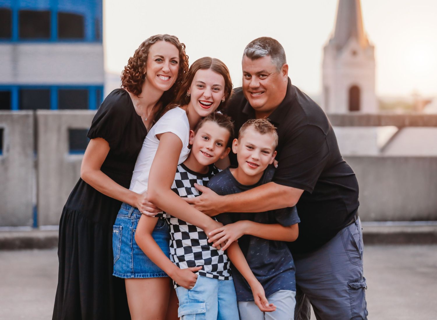 A group embraces and smiles together outdoors with a church steeple visible in the background.