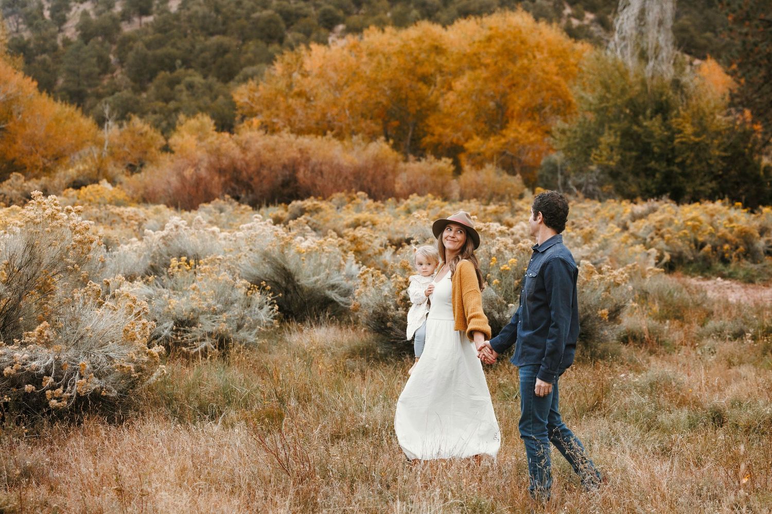 Young family with baby walks through golden autumn field with colorful fall foliage and rocky cliffs in background.