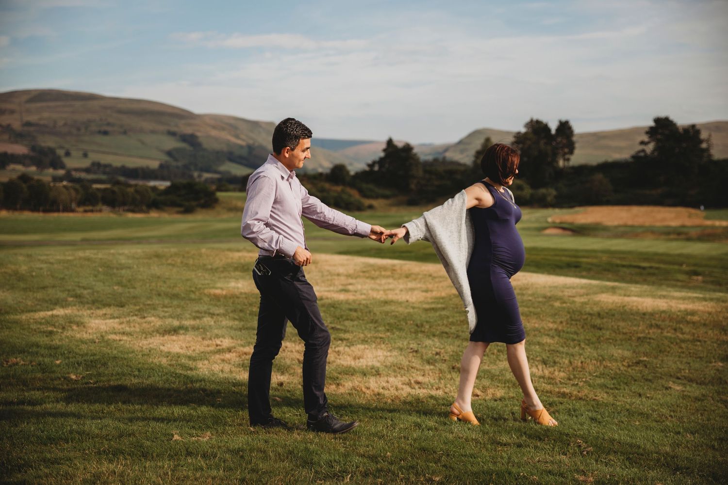A couple dances playfully in a grassy field with rolling hills and mountains in the background during golden hour.