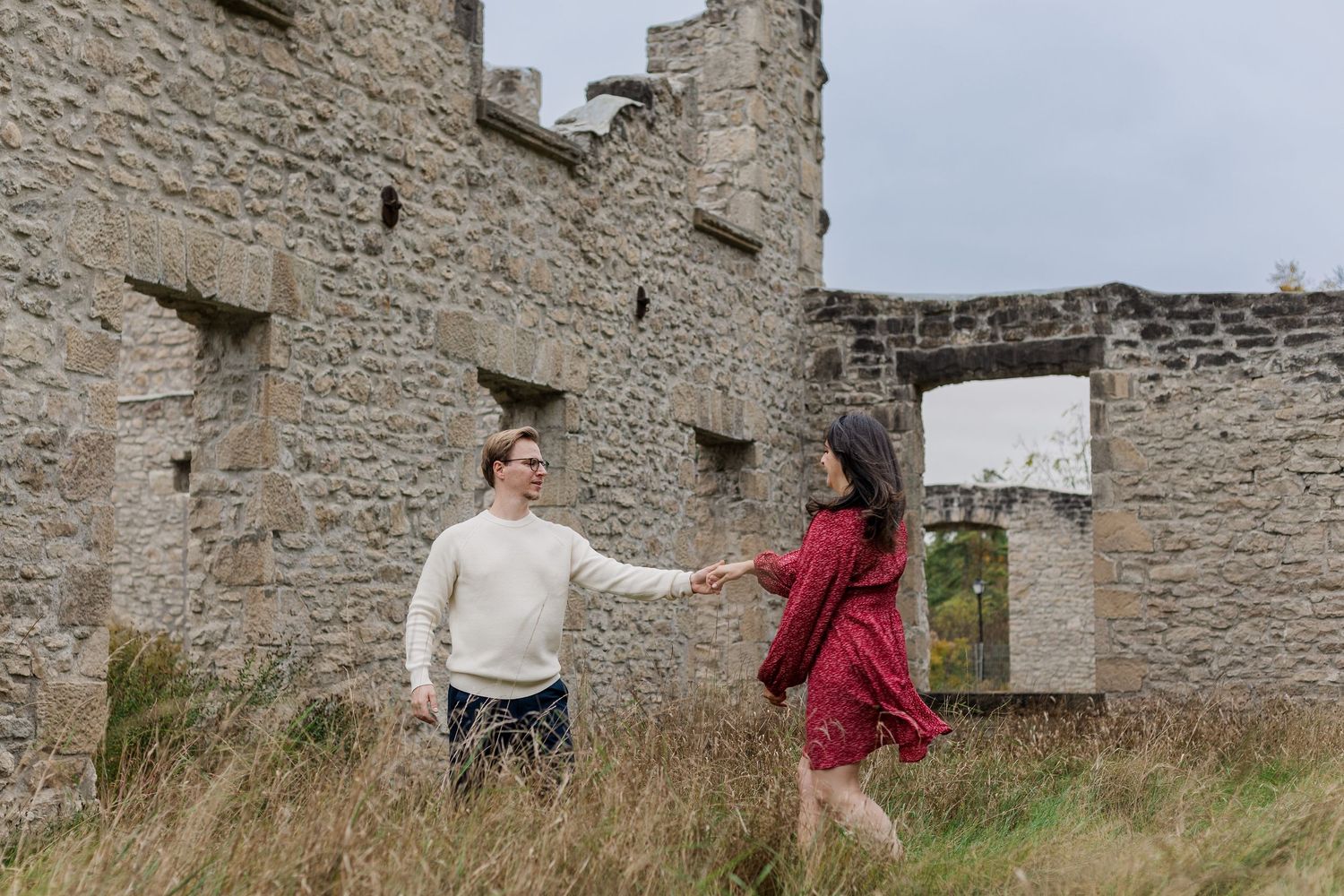 Two people explore ancient stone castle ruins together on an overcast day in a grassy field.
