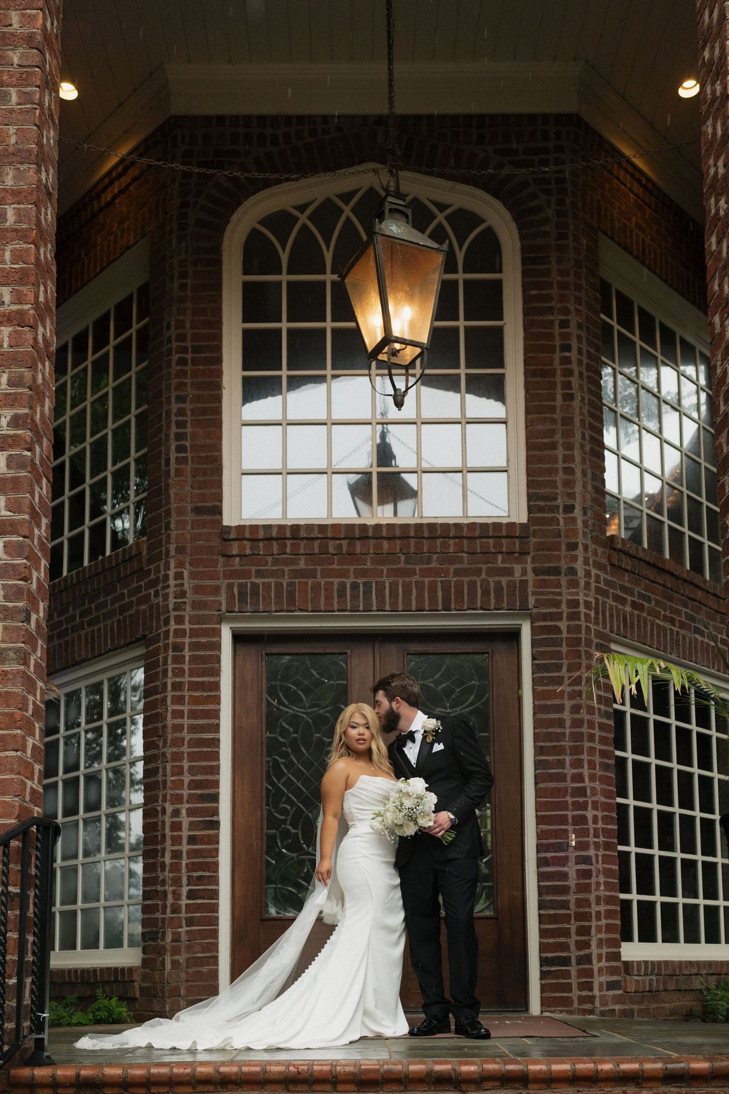 A romantic wedding portrait in front of a brick building with arched windows and glowing lantern.