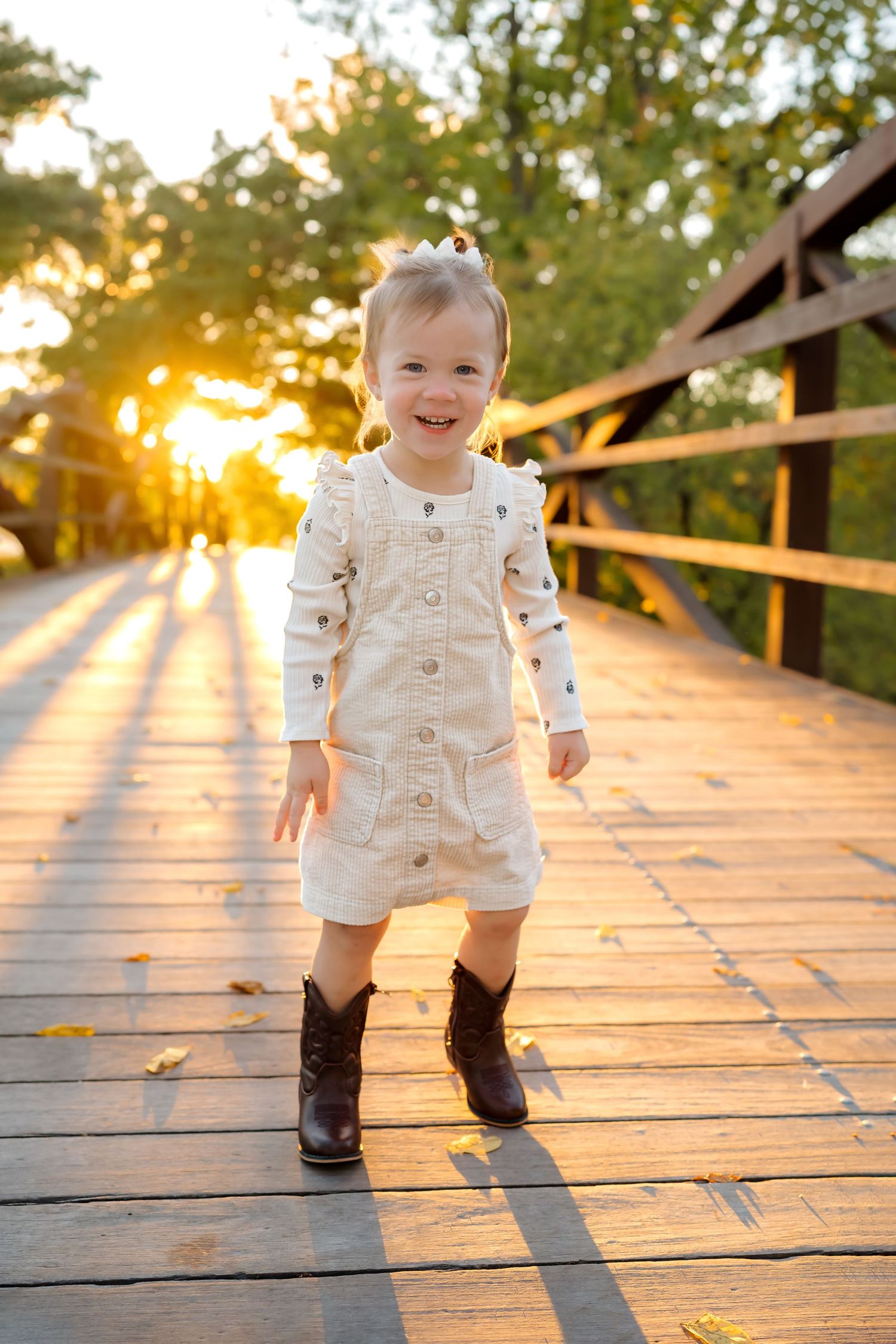 A toddler in a white romper and cowboy boots stands on a wooden bridge at sunset.