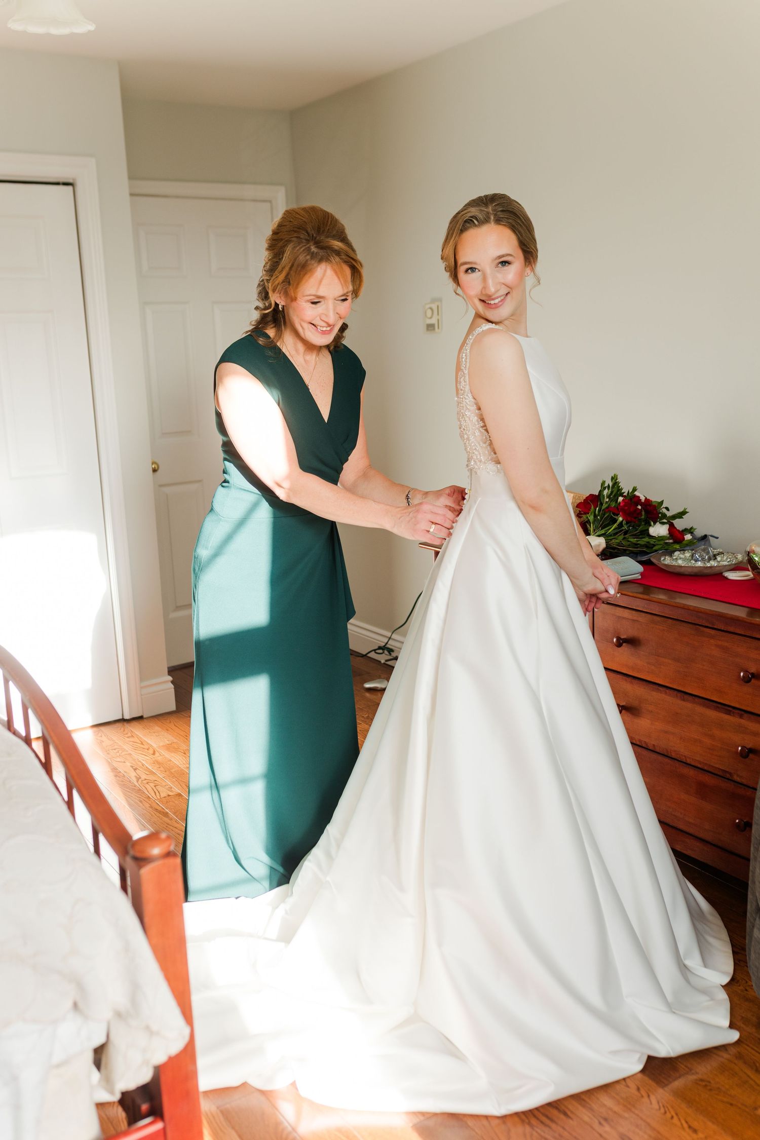 A person in a green dress helps adjust a white wedding gown in a sunlit room with wooden furniture.