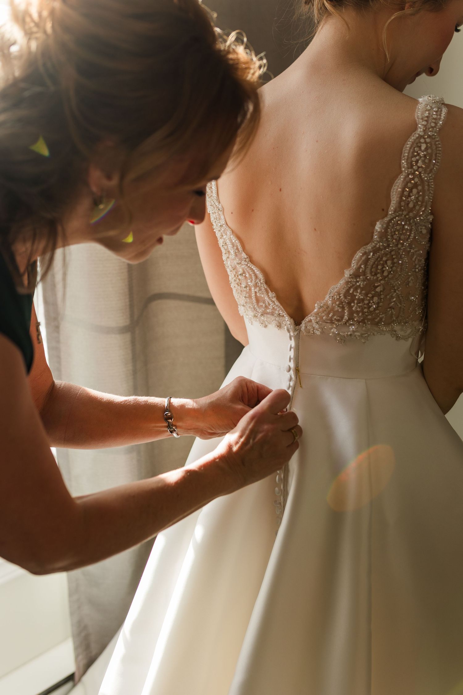 Close-up shots of hands fastening delicate buttons on the back of an ornate white wedding gown.