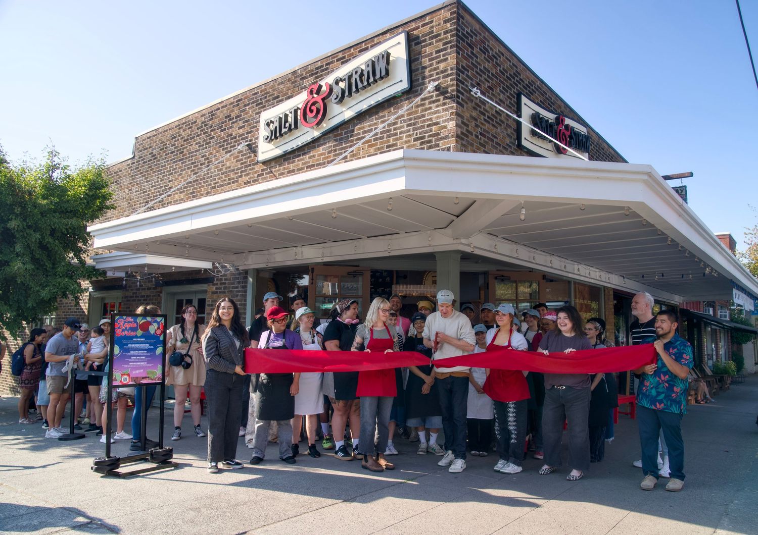 A group of people outside of the ice cream store, Salt and Straw, stand behind a red ribbon to announce the store's opening day.