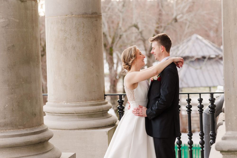 Romantic winter wedding couple sharing intimate moments between elegant stone columns at historic venue.