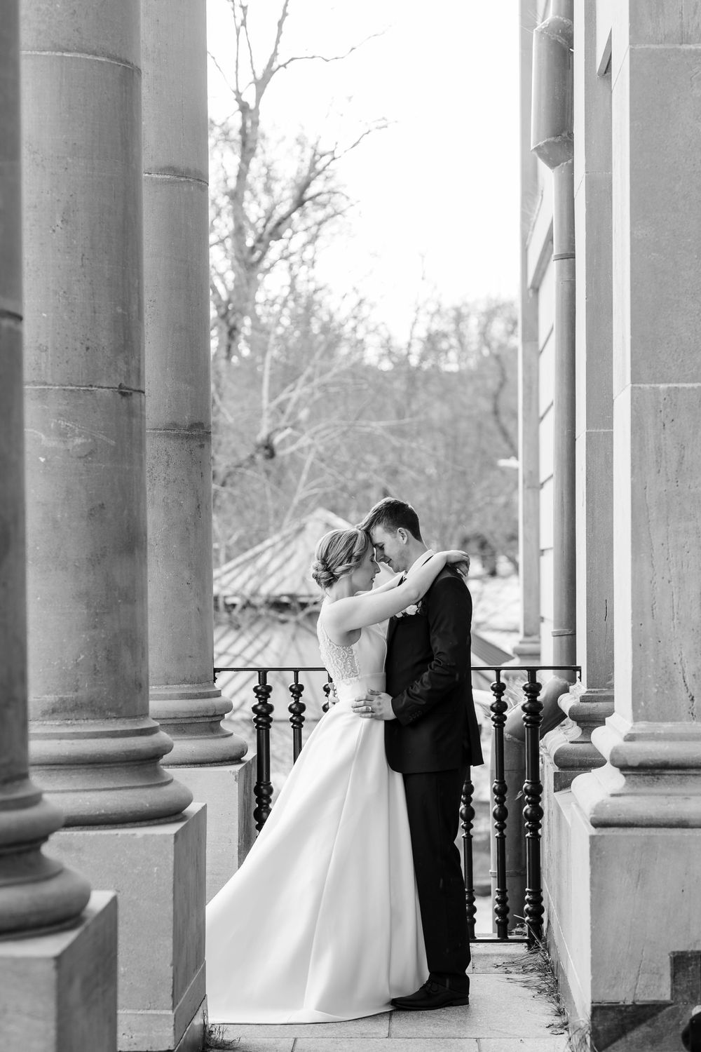 A romantic black and white wedding photo showing a couple embracing between elegant stone columns on a balcony.