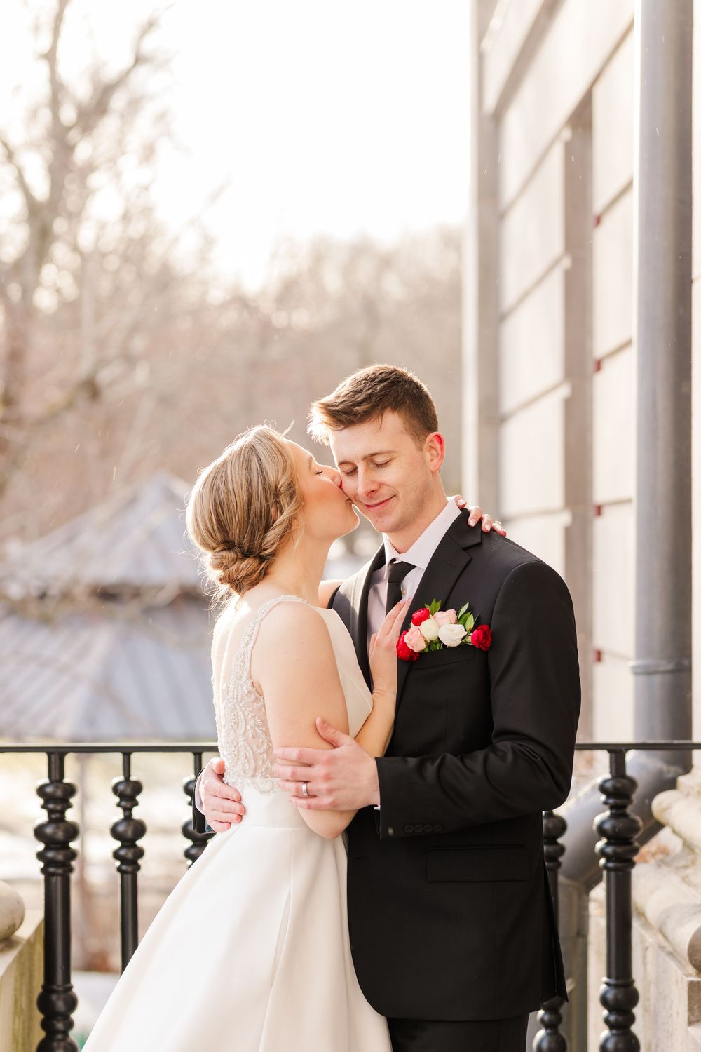 A romantic wedding couple embraces on a porch during sunset, with the bride in white and groom in classic black suit.