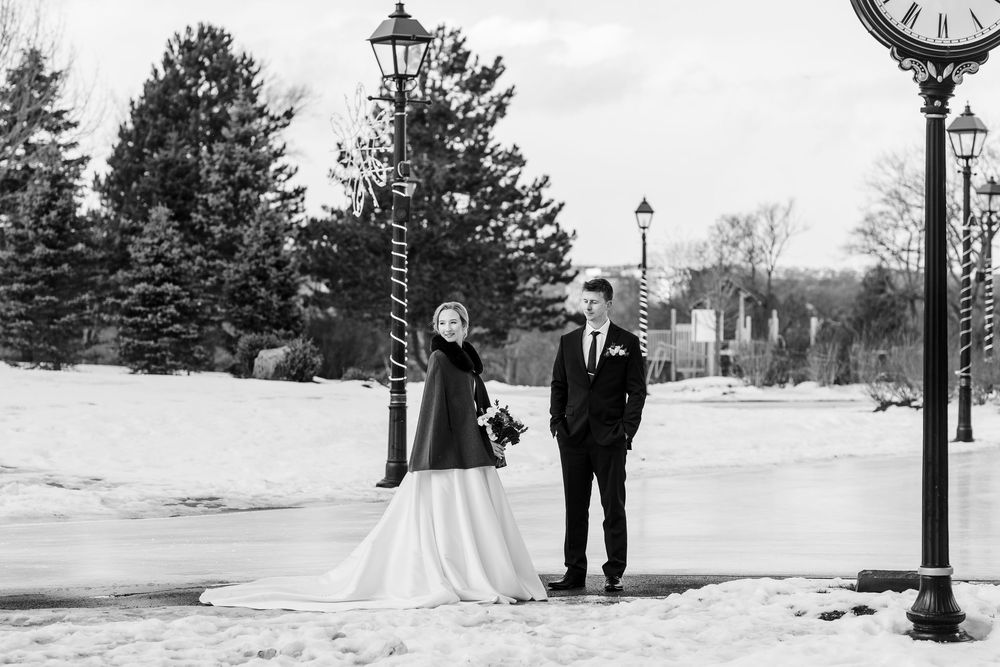 A winter wedding couple walks through snow covered grounds with vintage lamp posts and evergreen trees in the background.