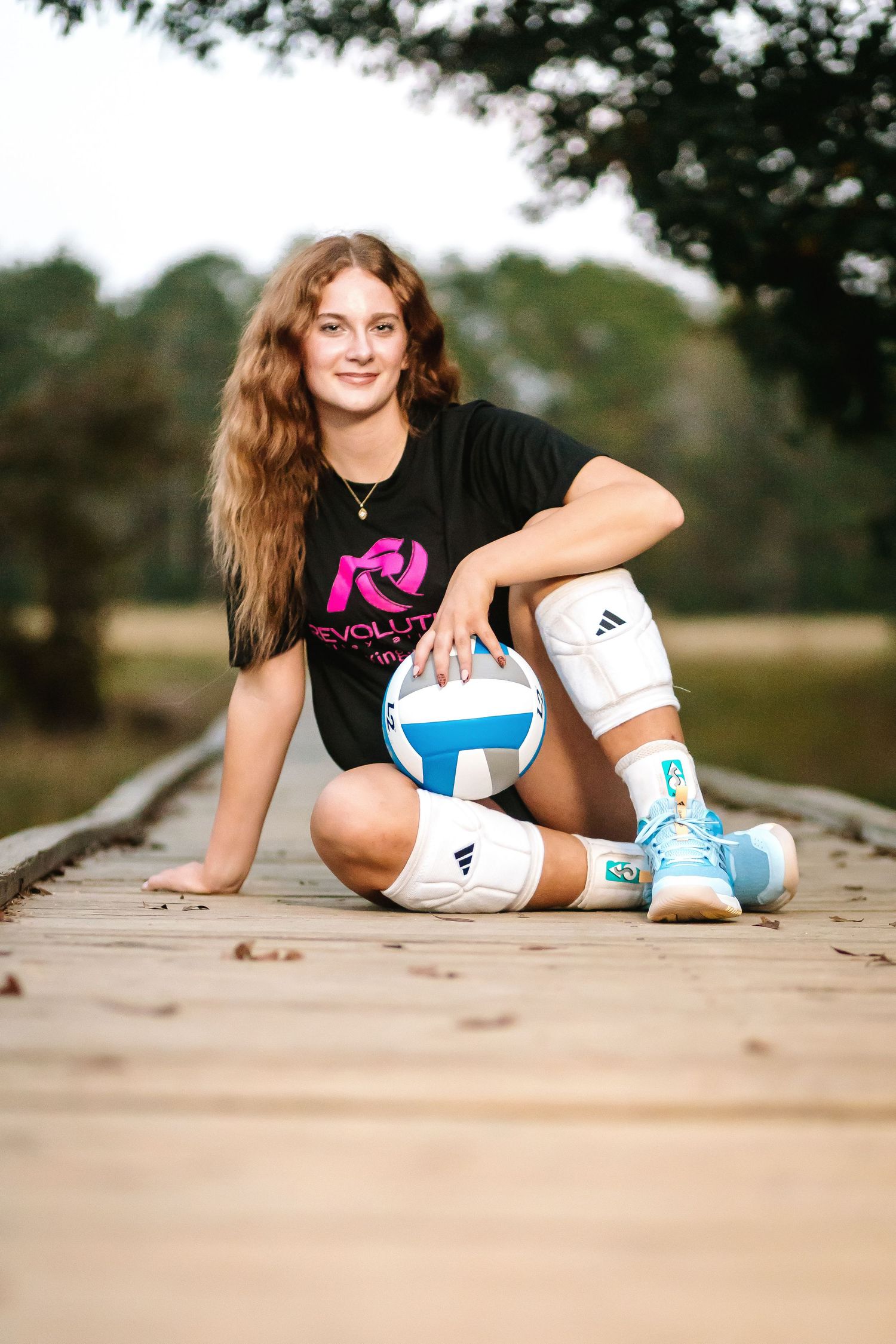 Volleyball player in black shirt and knee pads poses on wooden surface with ball in outdoor setting.
