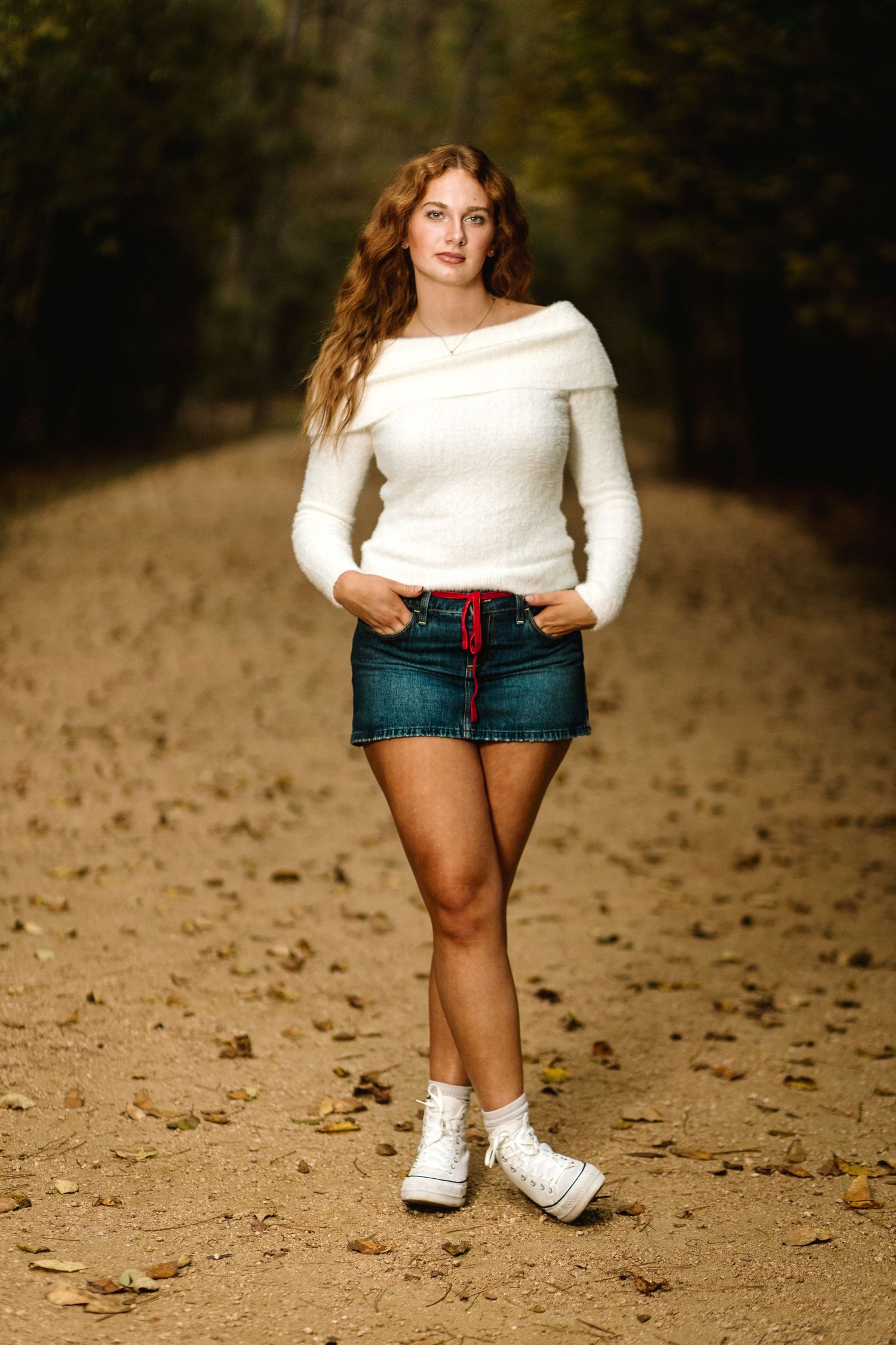 Person in white top and denim shorts standing on dirt path with autumn leaves scattered around.