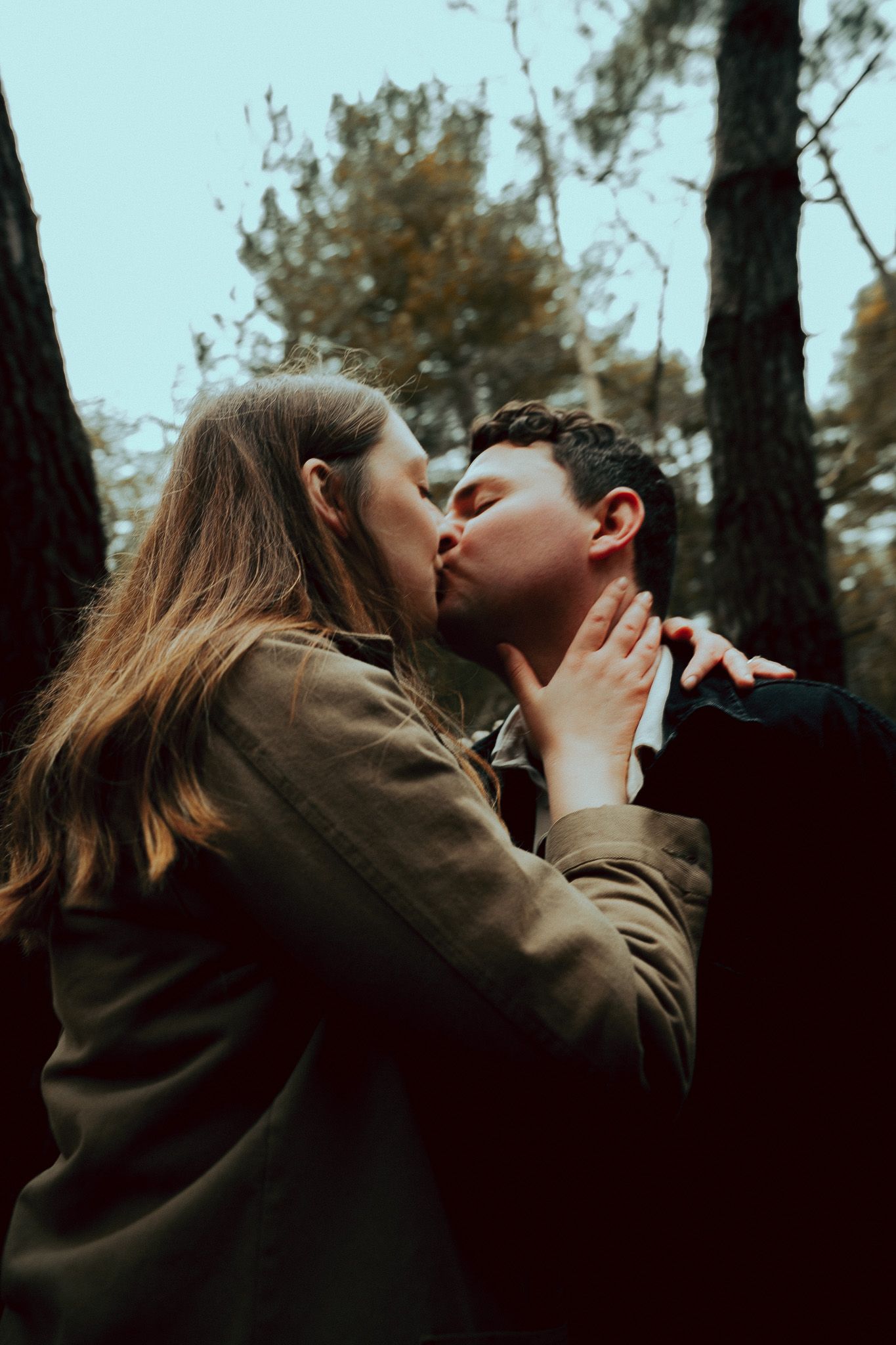 A couple shares a romantic kiss in a forest setting with pine trees visible in the background.