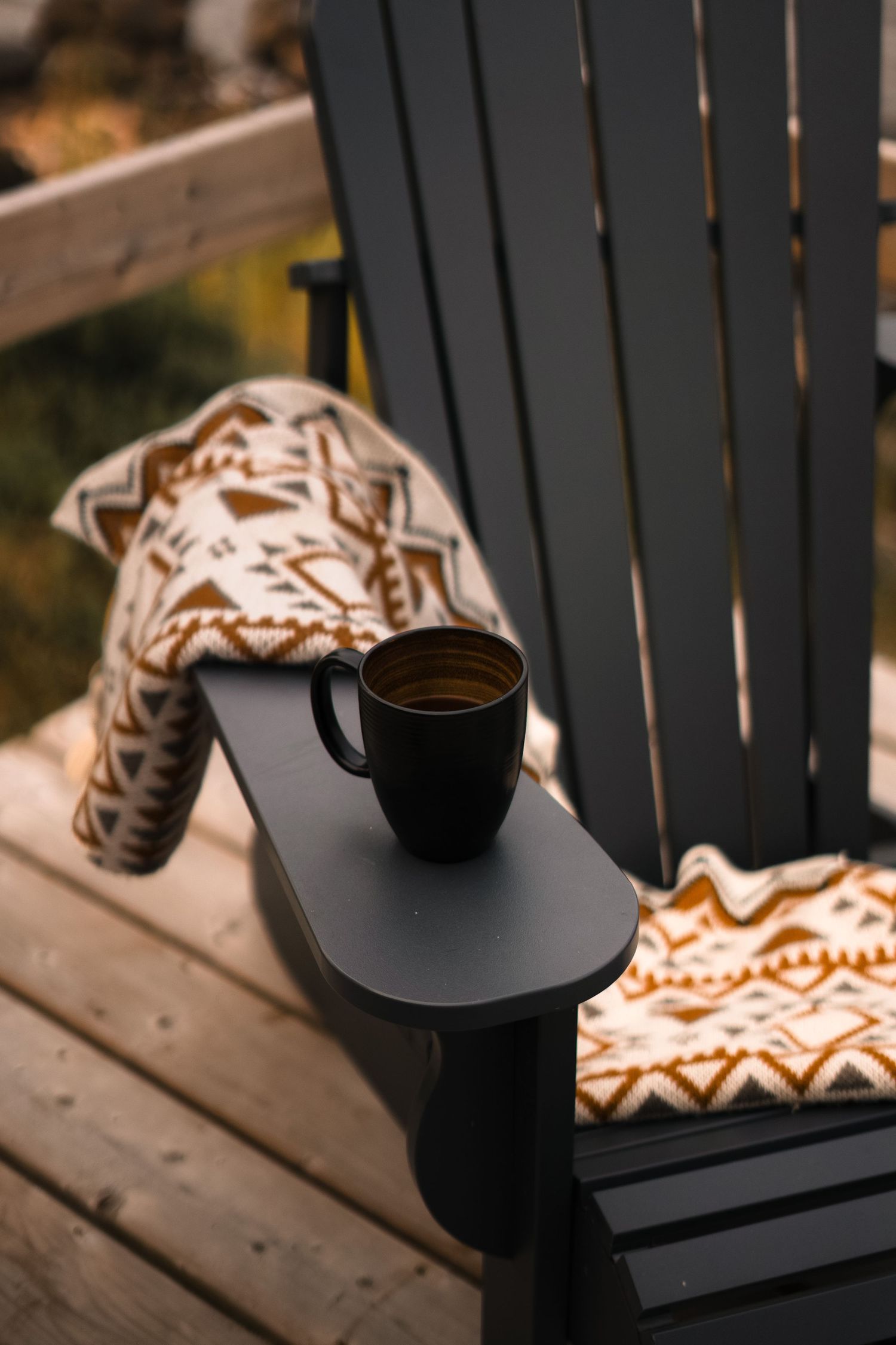 Black Adirondack chair with patterned blanket and coffee mug on wooden deck.