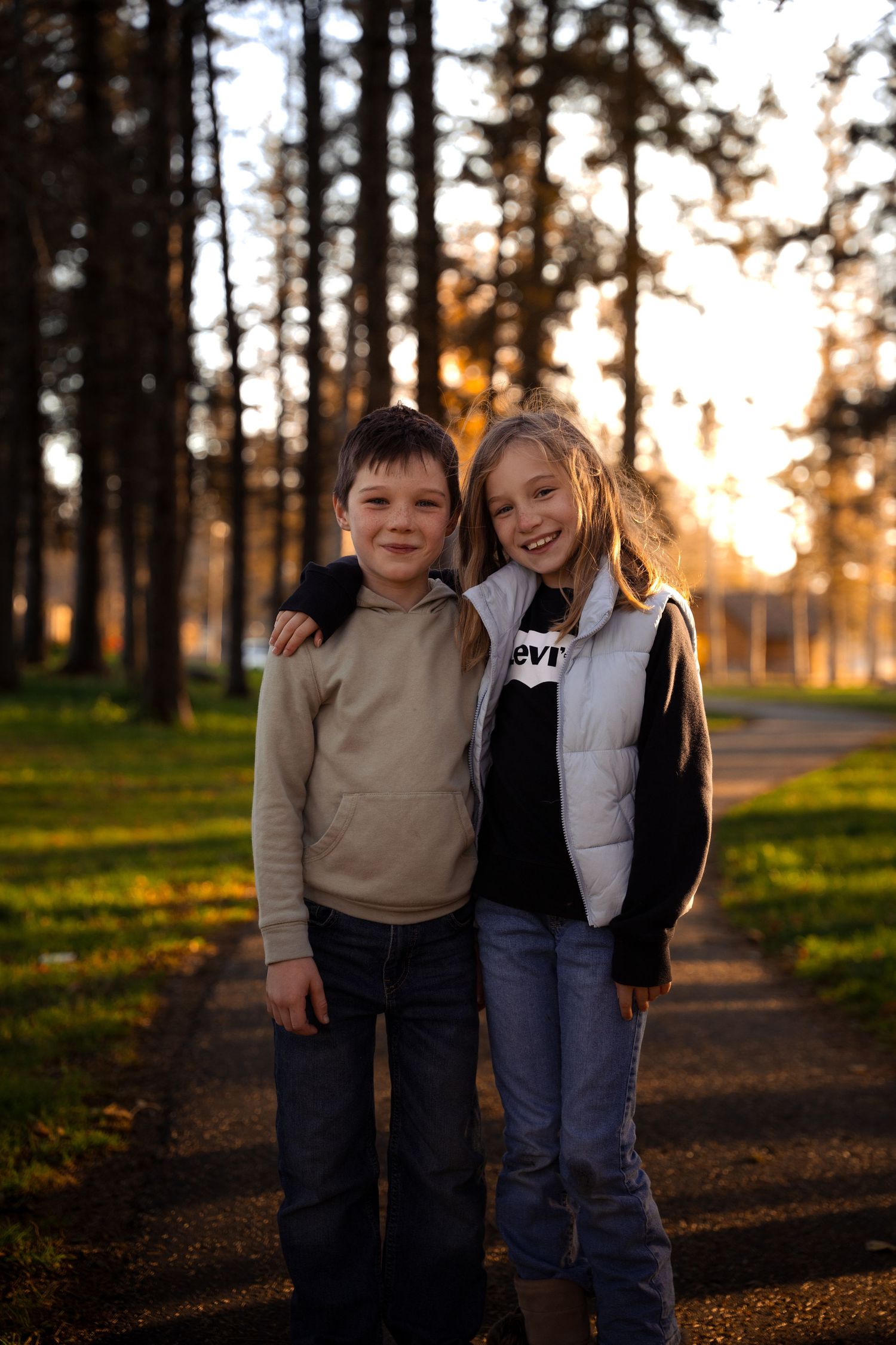 Two children stand together on a path in a forest during golden hour sunset.