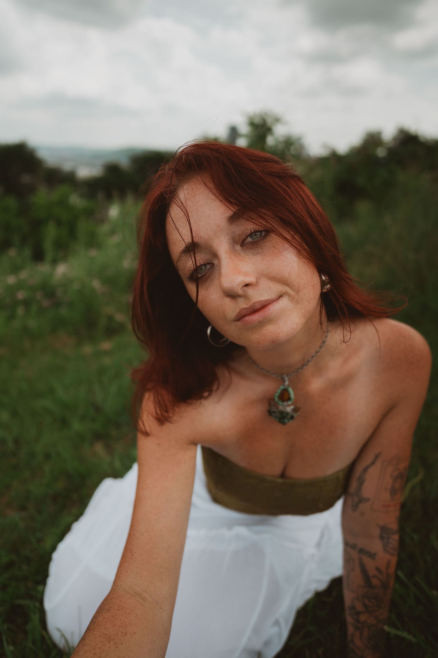 A person with red hair and tattoos poses outdoors in a white tank top against a natural green background.