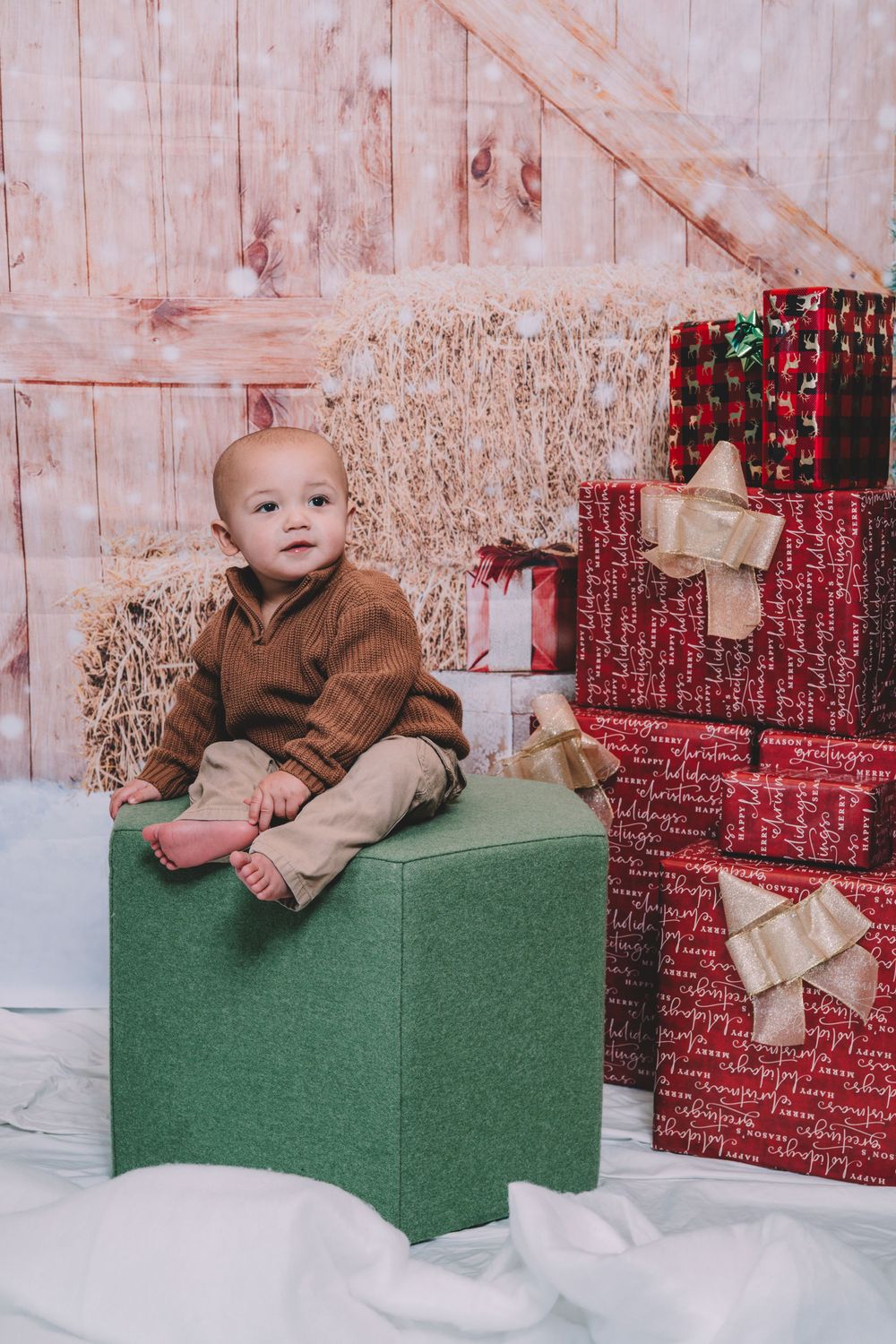 Baby in brown sweater sitting on green box with Christmas presents in background.