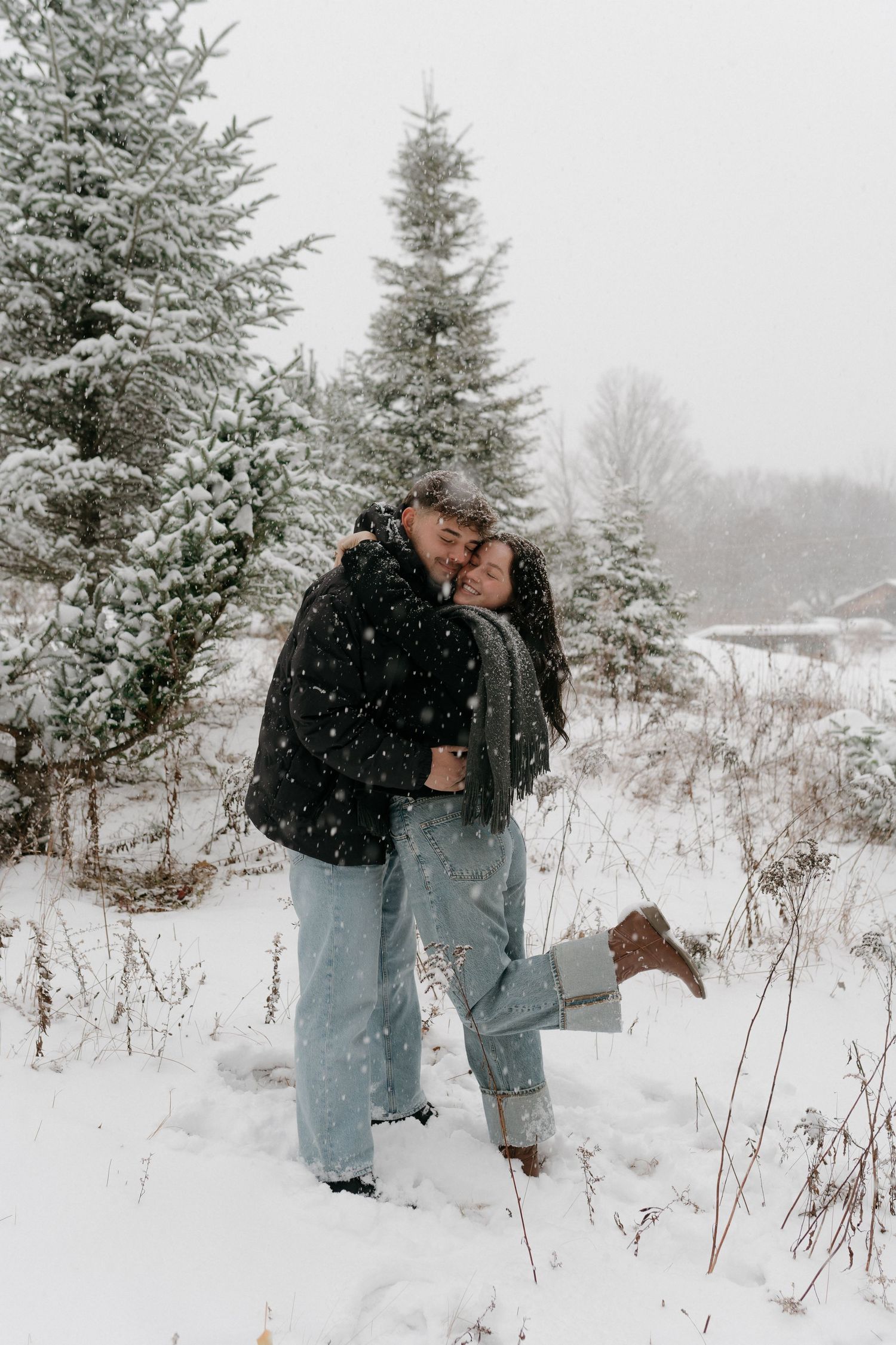 A couple embraces in a snowy winter landscape surrounded by snow-covered evergreen trees.