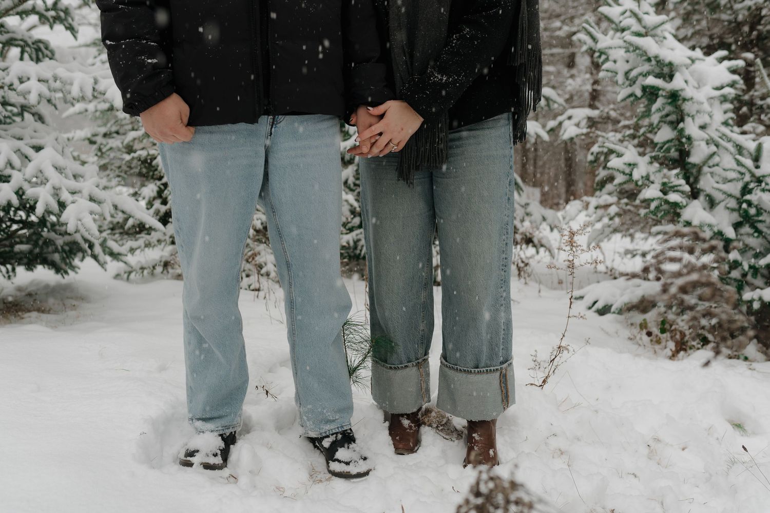 A couple in light blue jeans and brown shoes stands together in snowy winter scenery.